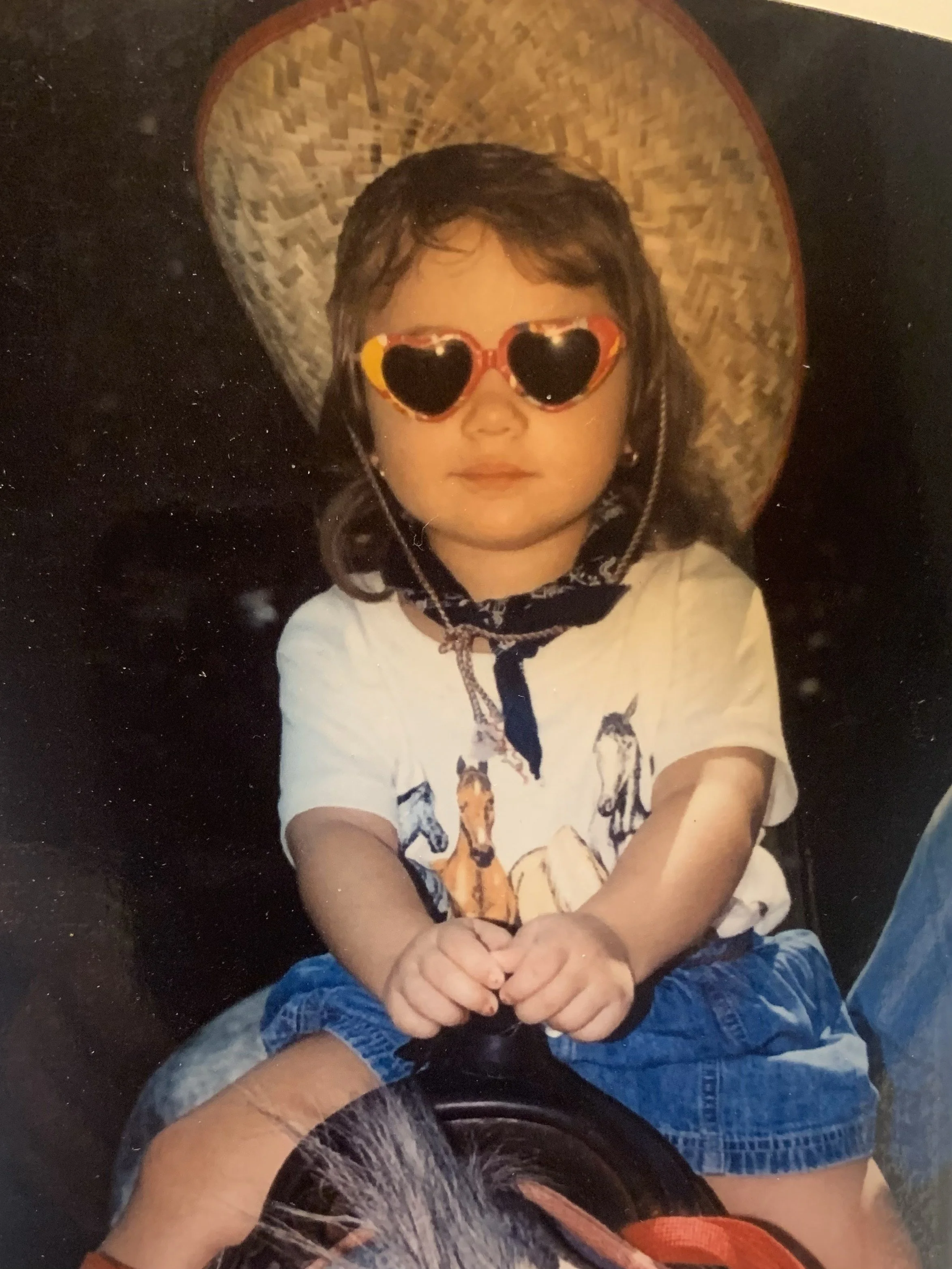 A young girl sitting in a stroller, wearing heart-shaped sunglasses, a white t-shirt with horse illustrations, blue shorts, and a black scarf, with a large wheat hat behind her.