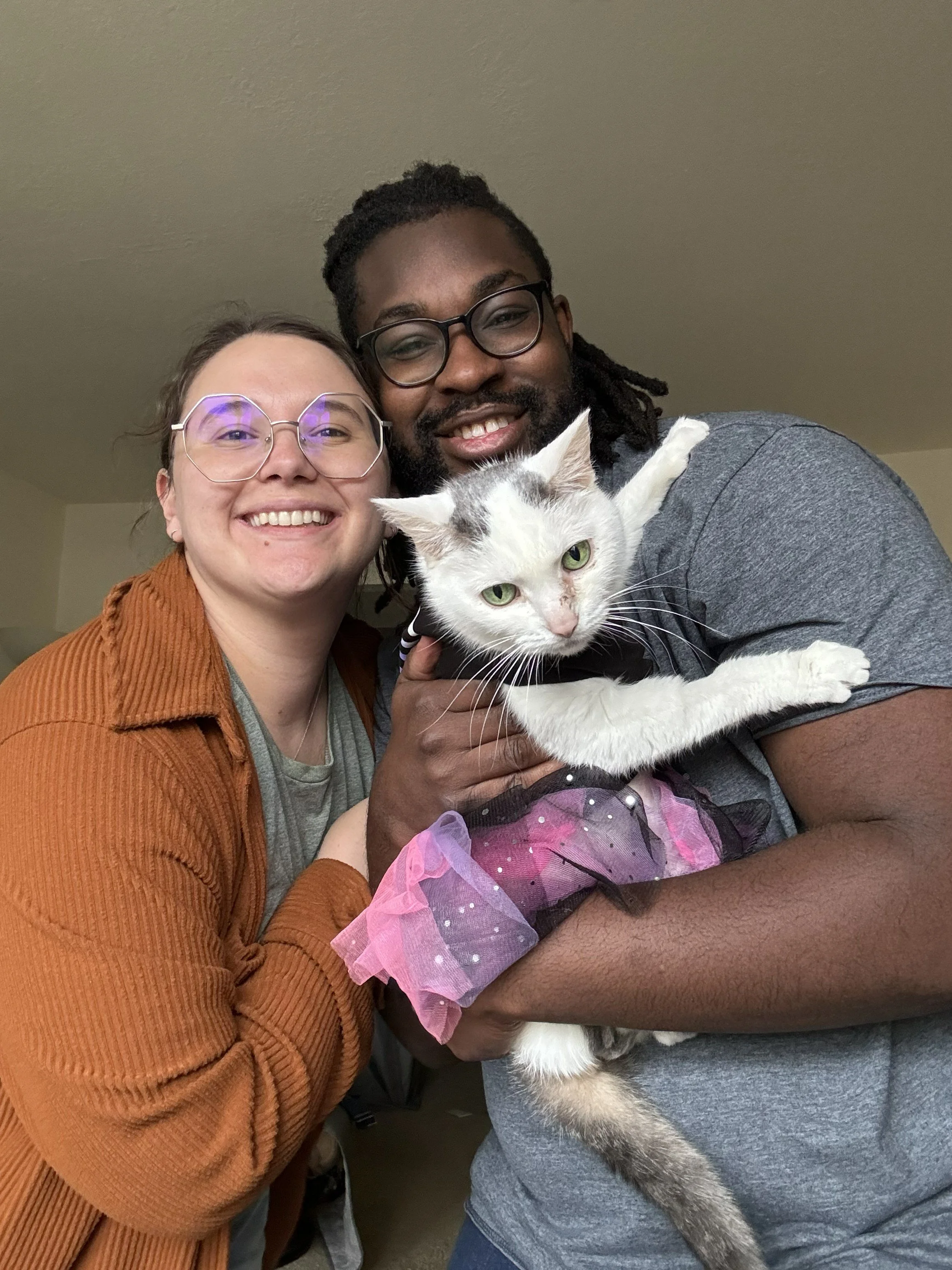 A happy couple with glasses holding a white cat with gray markings and a pink tutu, smiling at the camera indoors.