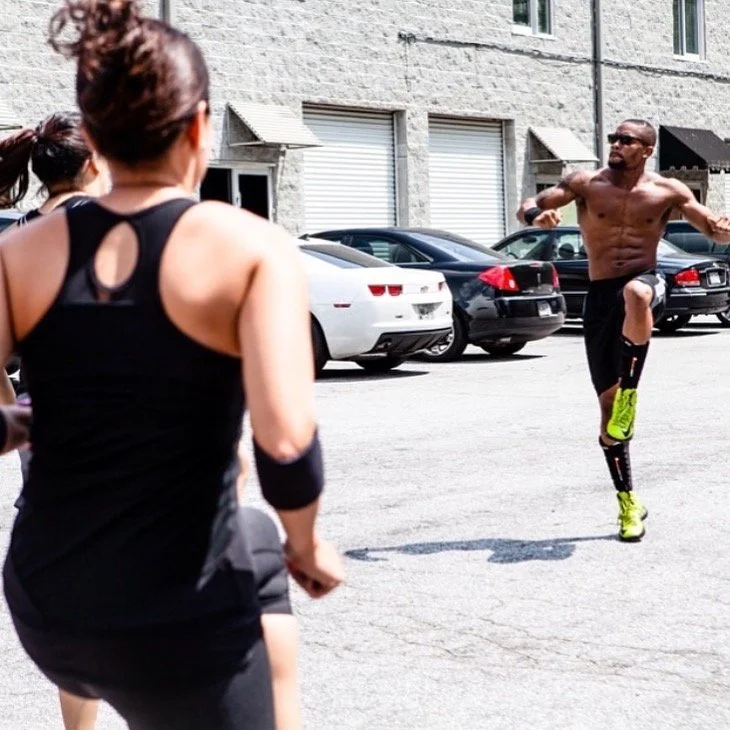 A group of runners, including a shirtless man with a prosthetic leg, warming up or exercising outdoors in front of a parking lot with parked cars and a building.