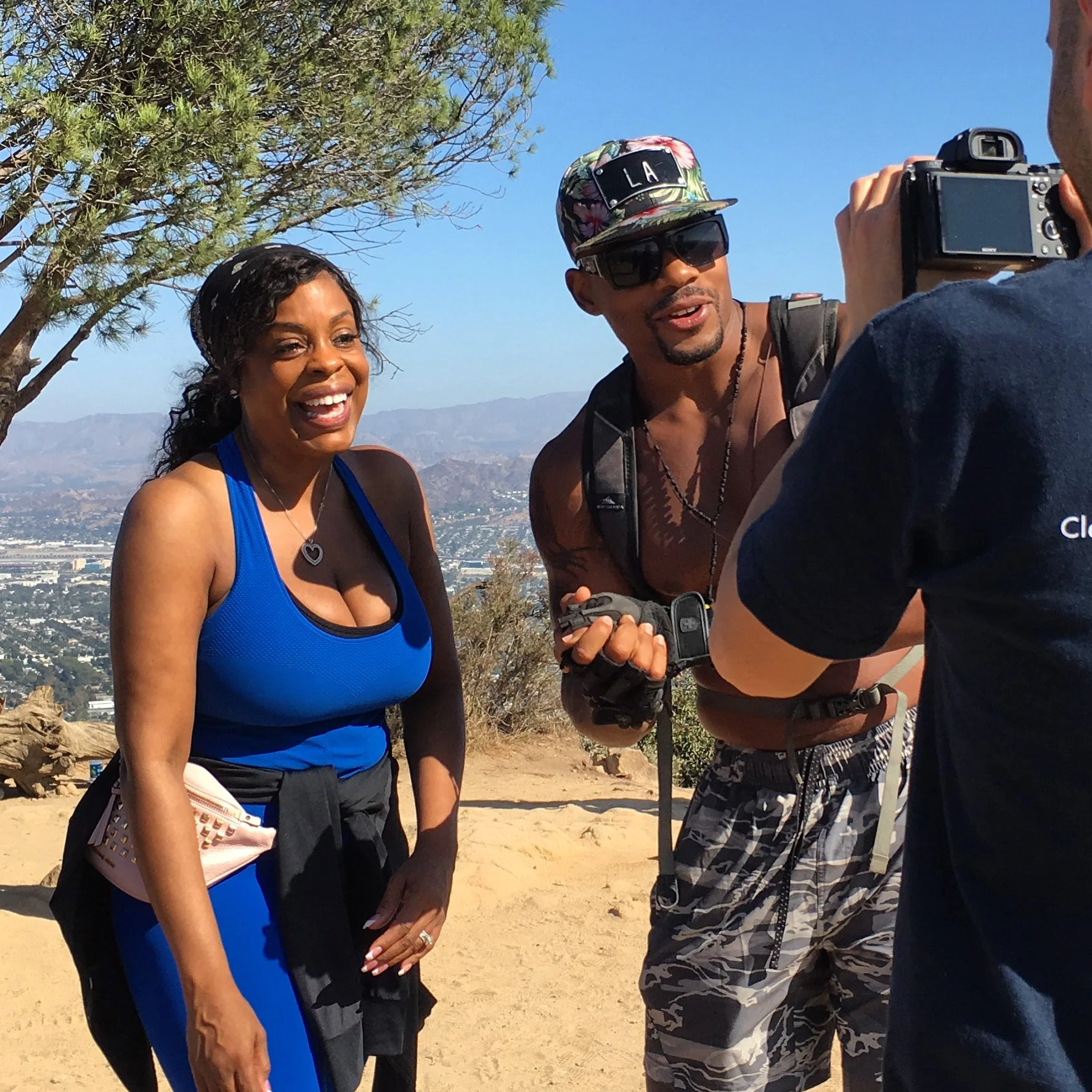 A woman in a blue workout outfit smiles while being filmed by a man with sunglasses and a shirt that says 'Cl', in a desert-like outdoor setting with mountains and a city in the background.