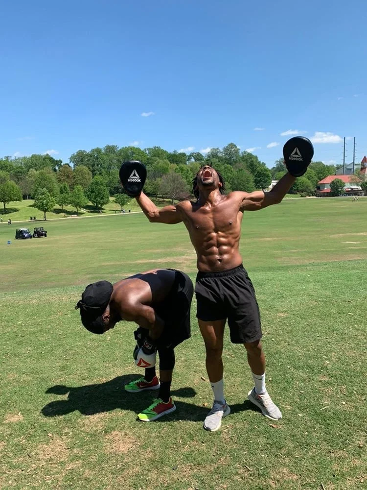 Two men in athletic clothing on a grassy field under a clear blue sky. One man, shirtless and barefoot, stands with arms raised holding weight plates and appears to be celebrating. The other man is bent over, adjusting his shoe.