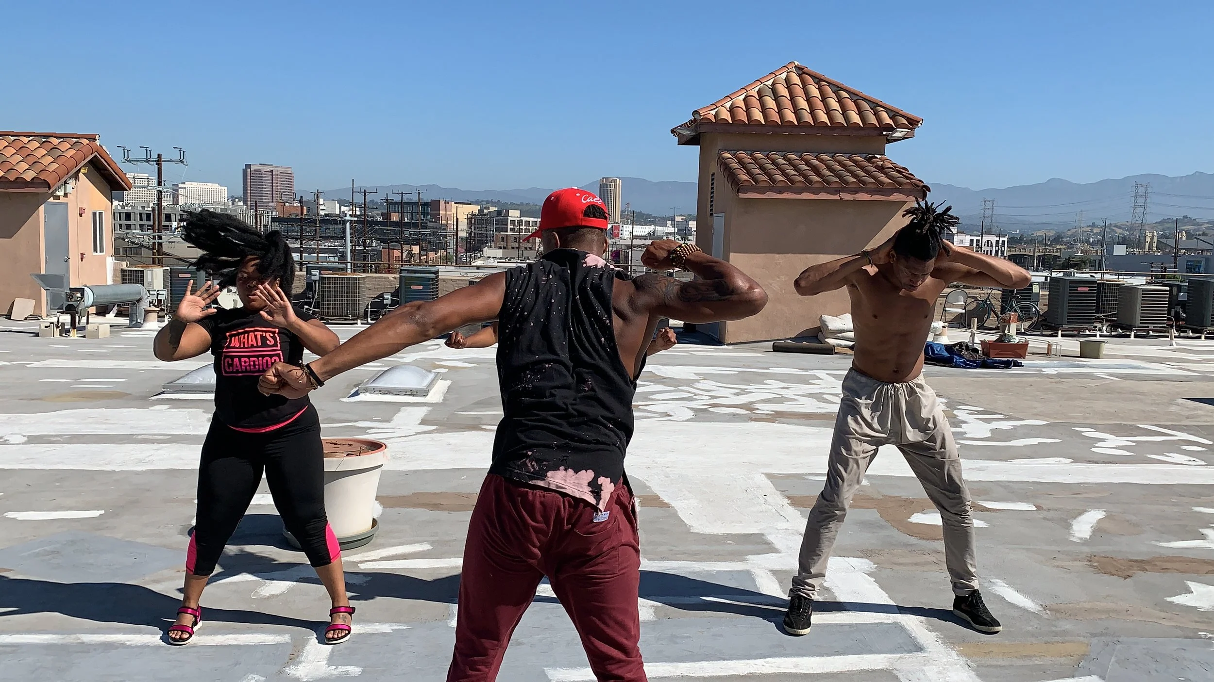 Three people dancing on a city rooftop with mountains in the background, two men and one woman, one man shirtless, the woman wearing black clothes, and the third man wearing a black tank top and red cap.