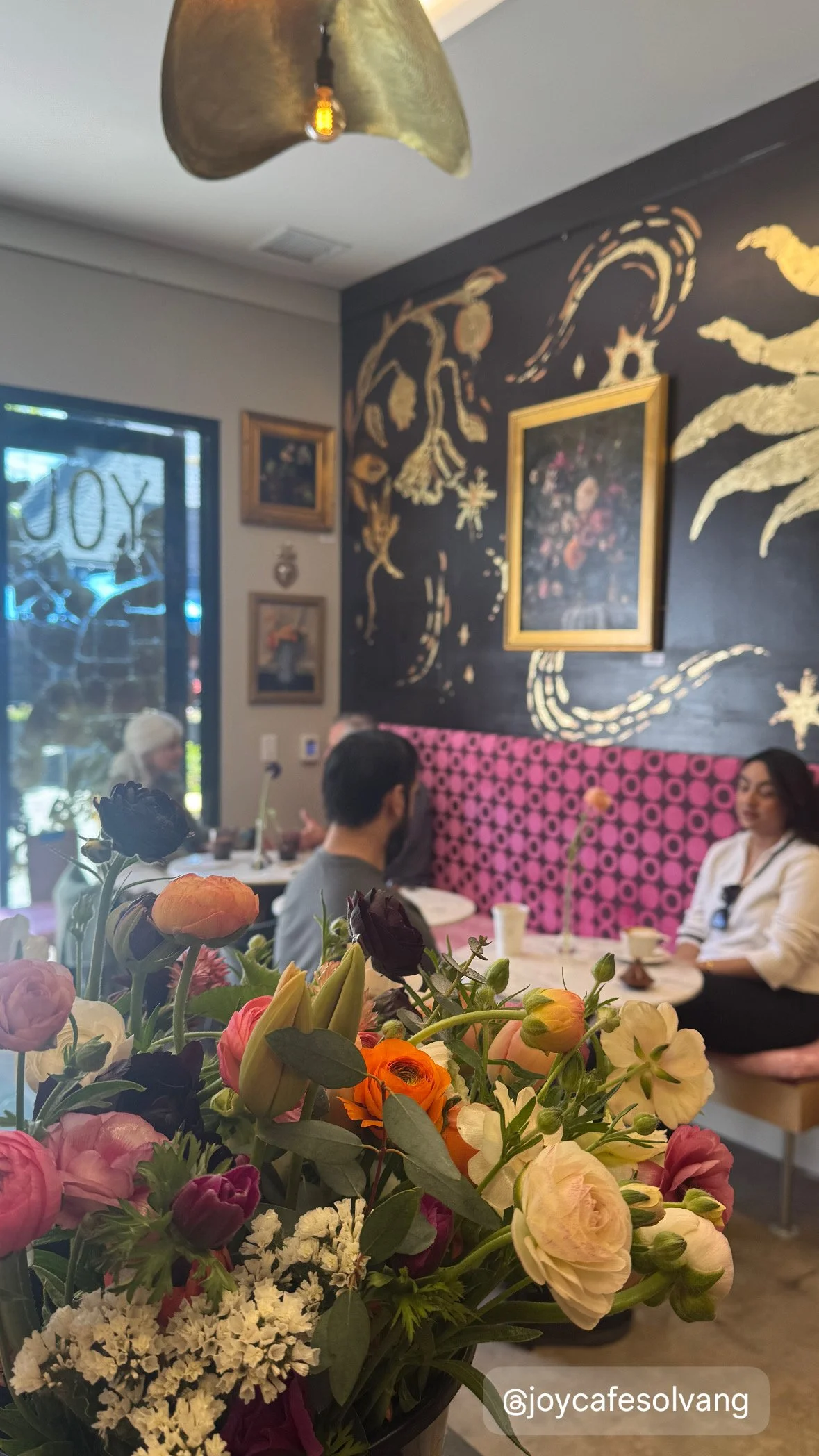 A close-up of a bouquet of colorful flowers on a table inside a cafe, with people dining in the background and framed artwork on the wall.