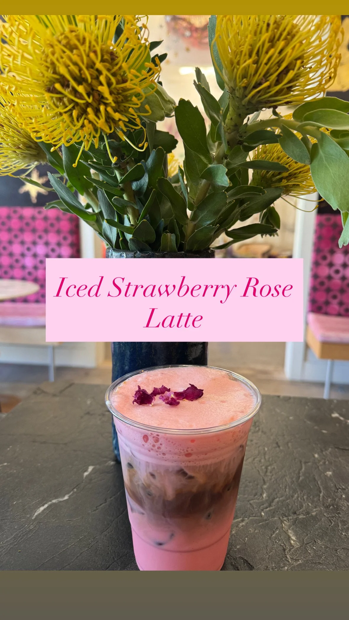 Pink iced strawberry rose latte in a clear glass with rose petals on top, positioned in front of a vase with yellow flowers and a pink sign with text.