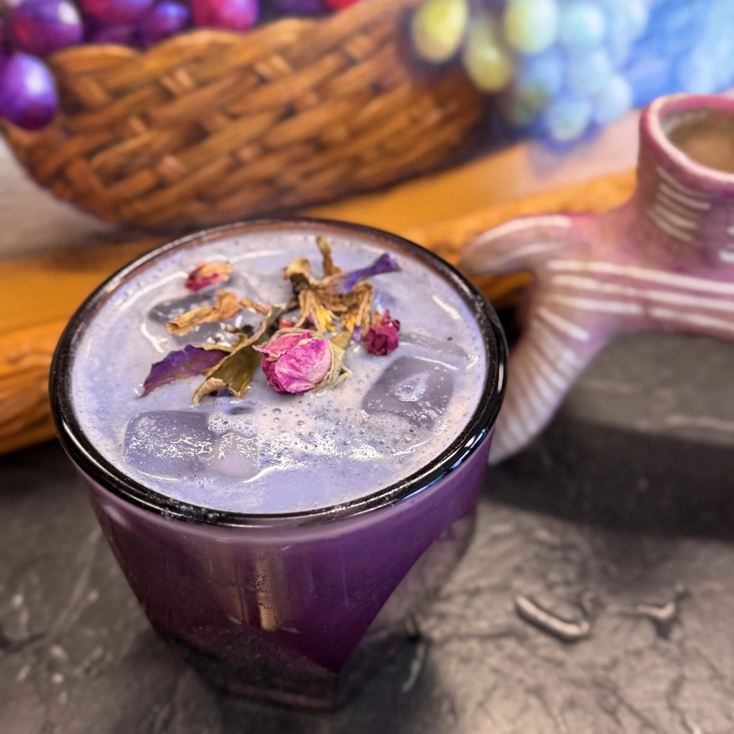 Close-up of a purple ceramic mug with a beverage inside, on a dark surface, with a blurred background of a colorful basket and another mug.