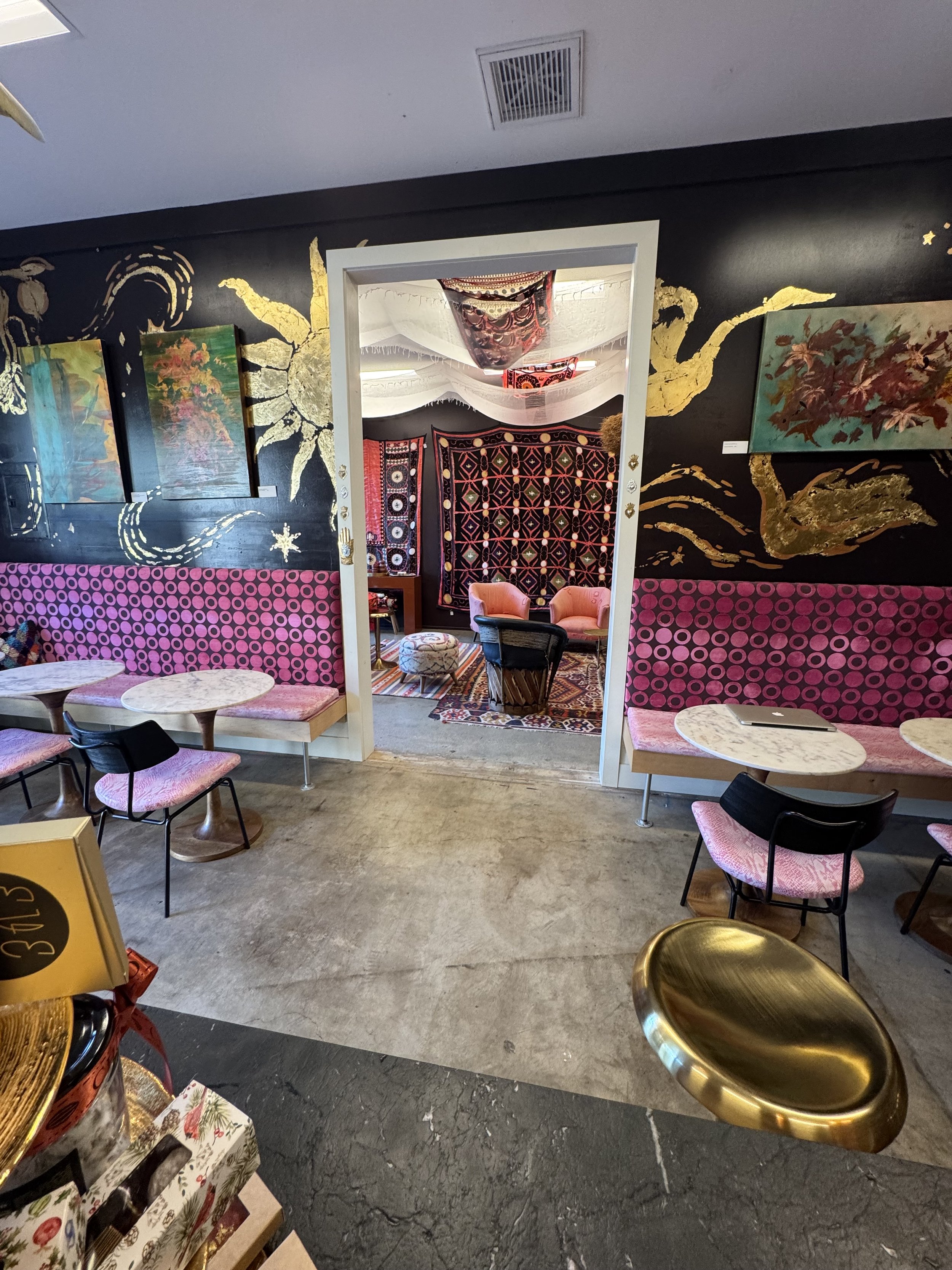 Interior of a colorful cafe or art space with patterned walls, paintings, pink seating, and tables. A mirror on the wall reflects more seating and decorations.