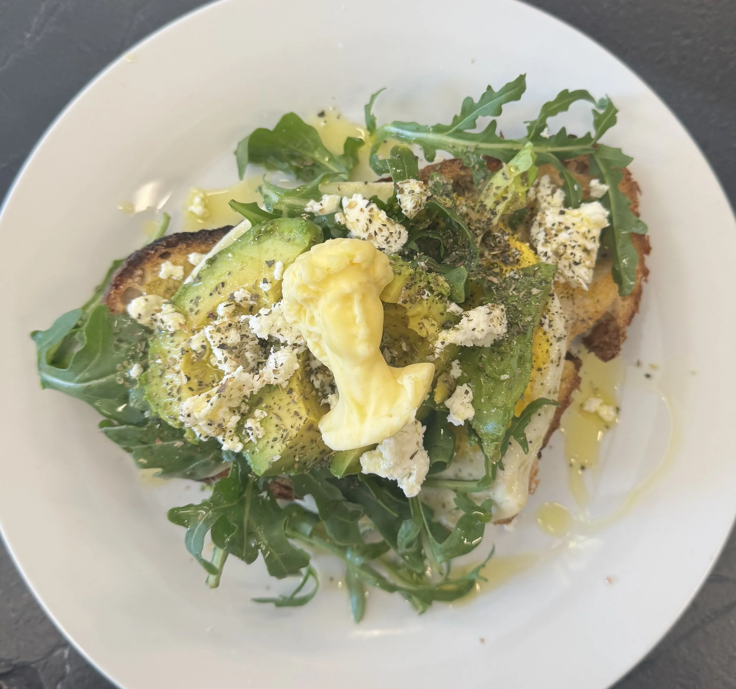 A plate with a slice of toasted bread topped with mashed avocado, arugula, crumbled cheese, and black pepper.
