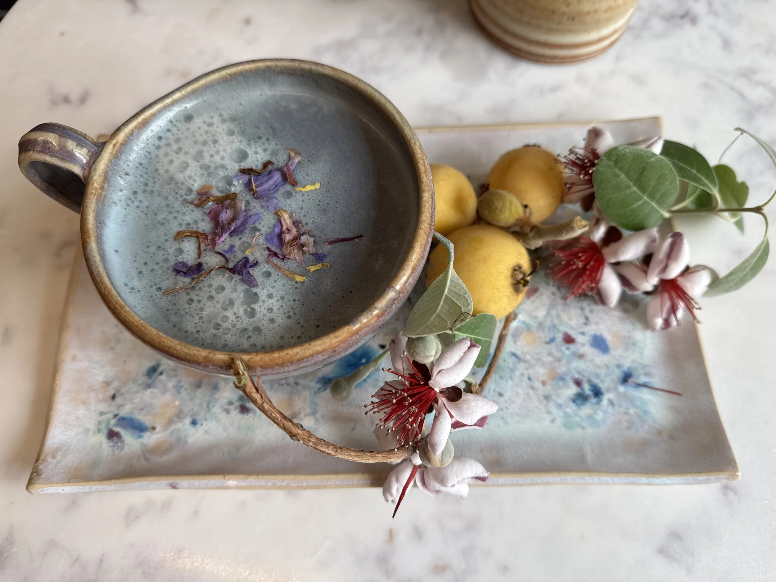 A ceramic mug filled with a frothy blue-gray beverage, topped with small purple and yellow flower petals, next to a white tray holding yellow fruits and green leaves, all on a marble surface.