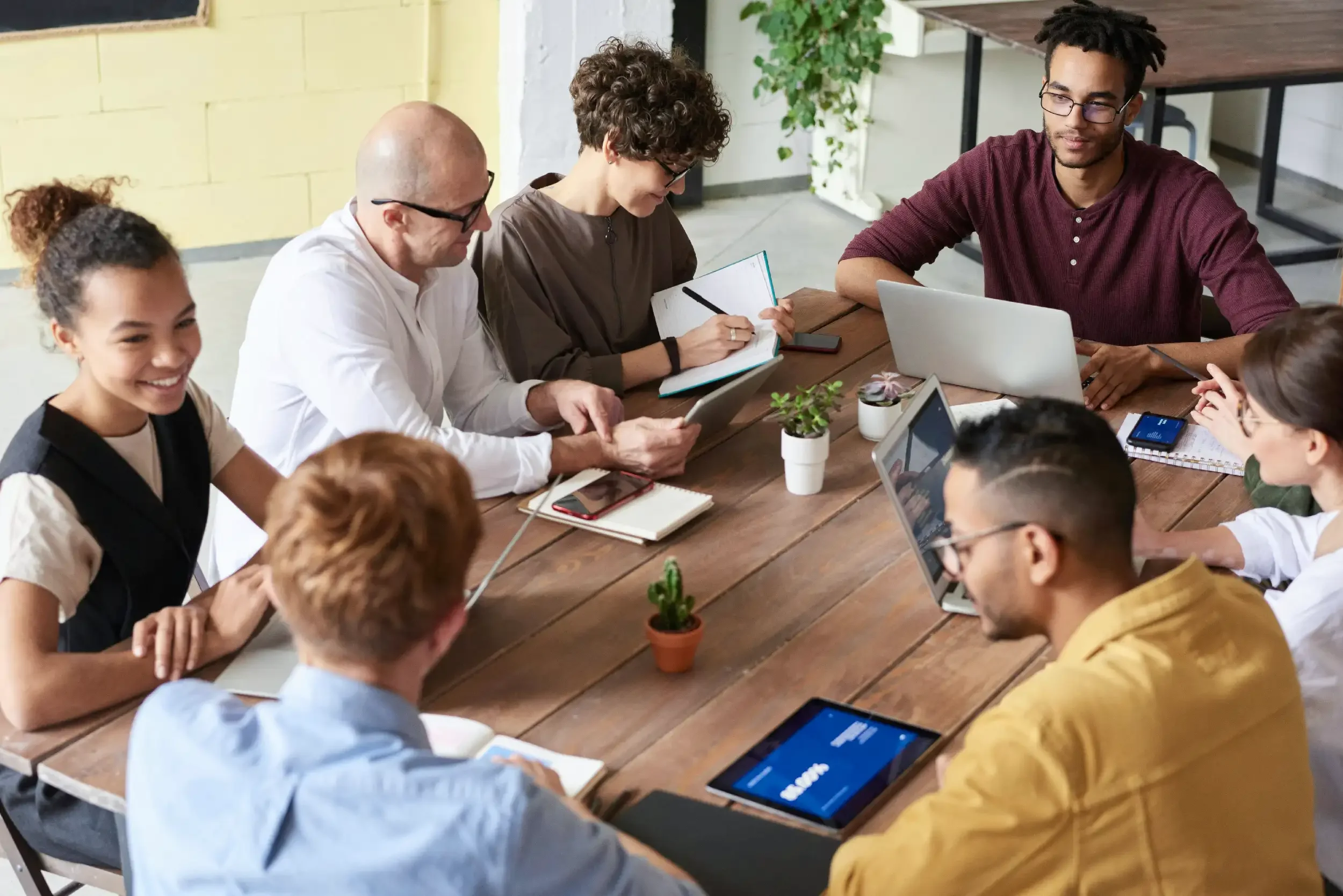 A diverse group of eight people sitting around a wooden table in a meeting room, engaged in discussion with laptops, tablets, notebooks, and smartphones.