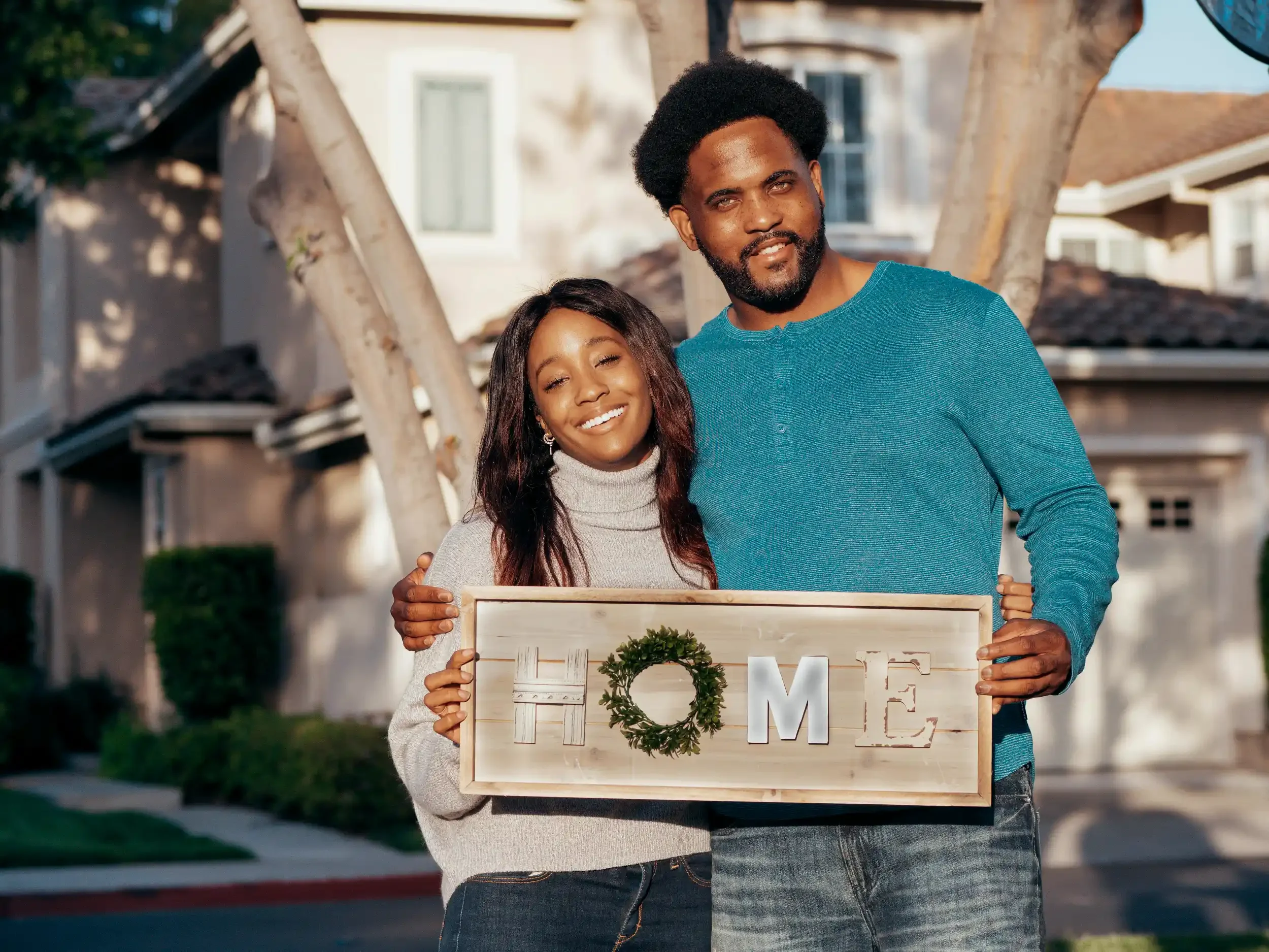 A couple standing outside holding a wooden sign that spells 'HOME' with a small wreath forming the letter 'O'. They are smiling and appear happy.