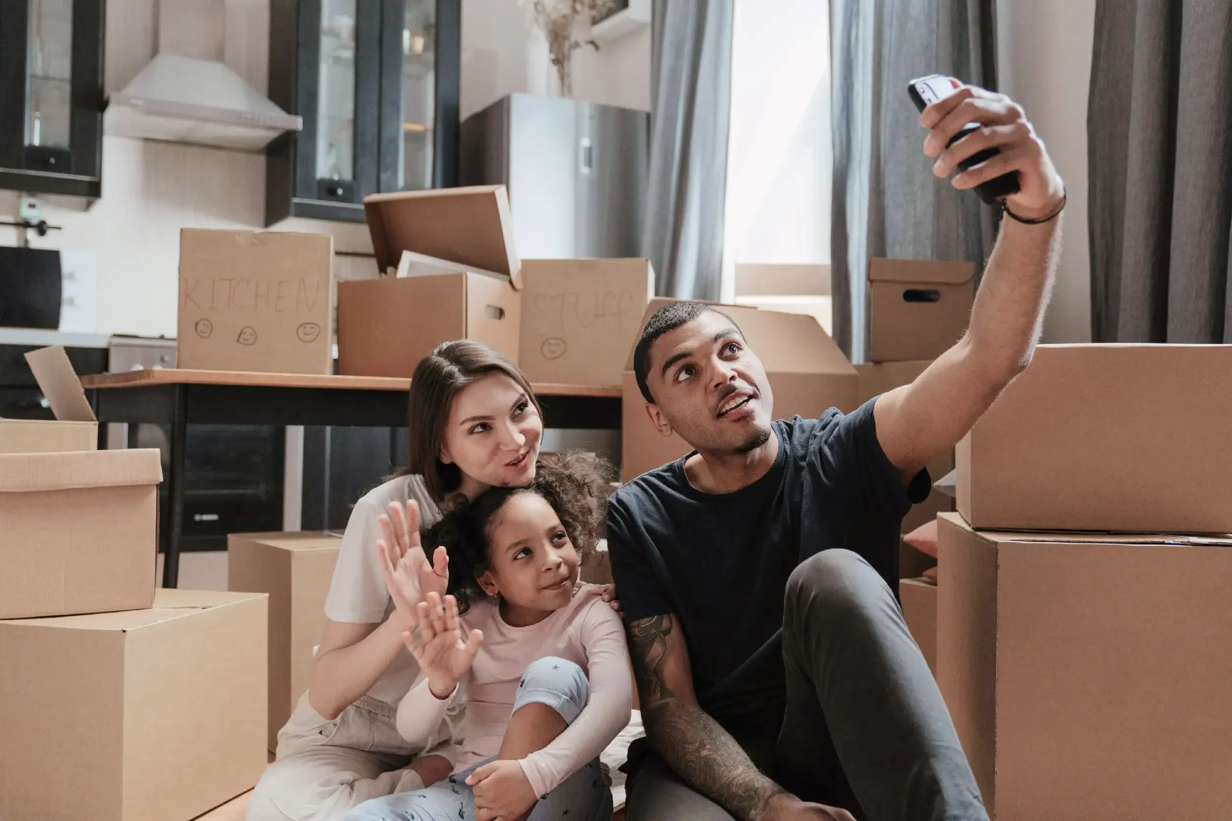 A family of three, two young girls and a man, sit among stacked moving boxes in a room, taking a selfie together.