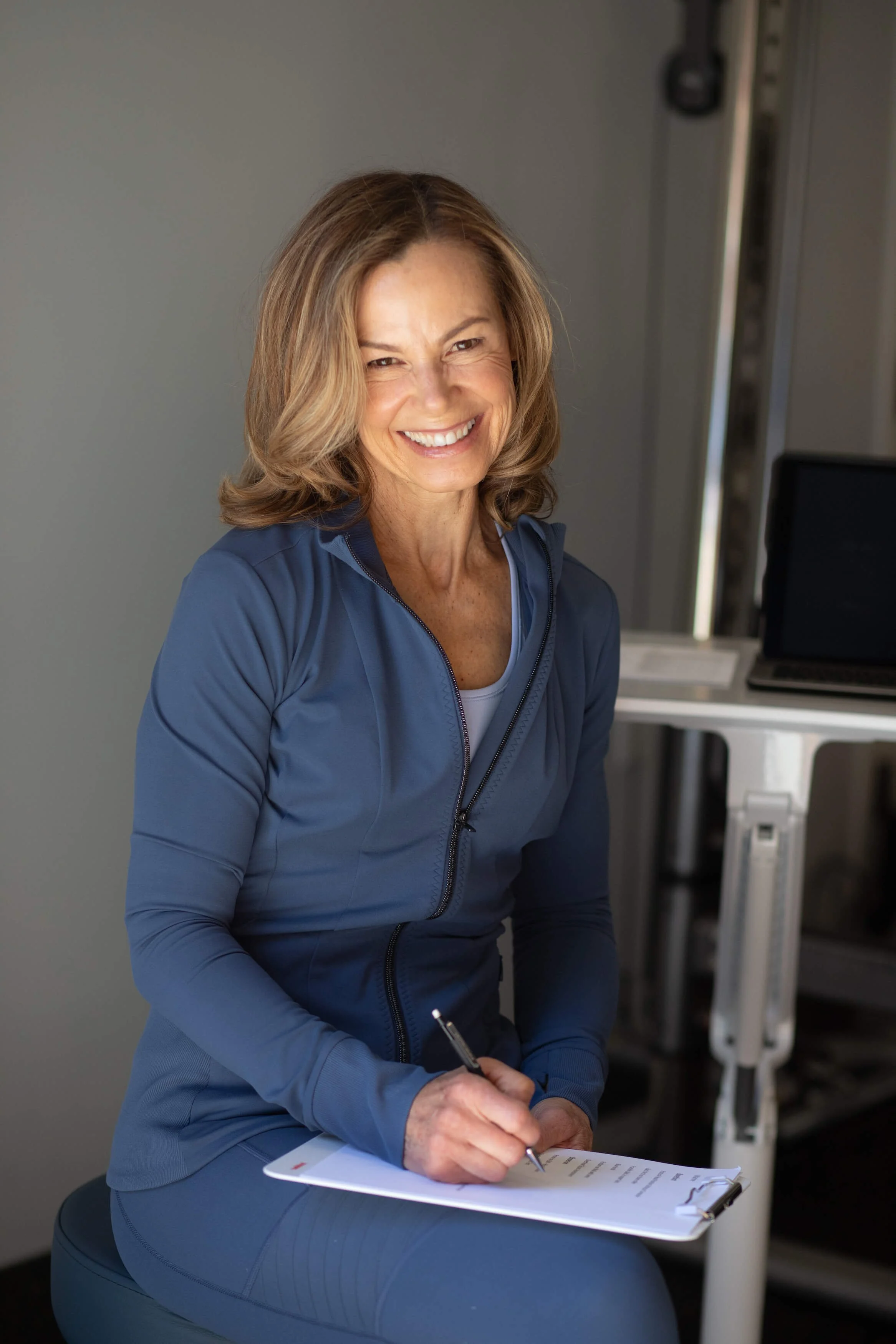 A smiling woman in athletic clothing sitting on a stool with a notepad and pen, in a studio setting with a gray background and a laptop on a cart.