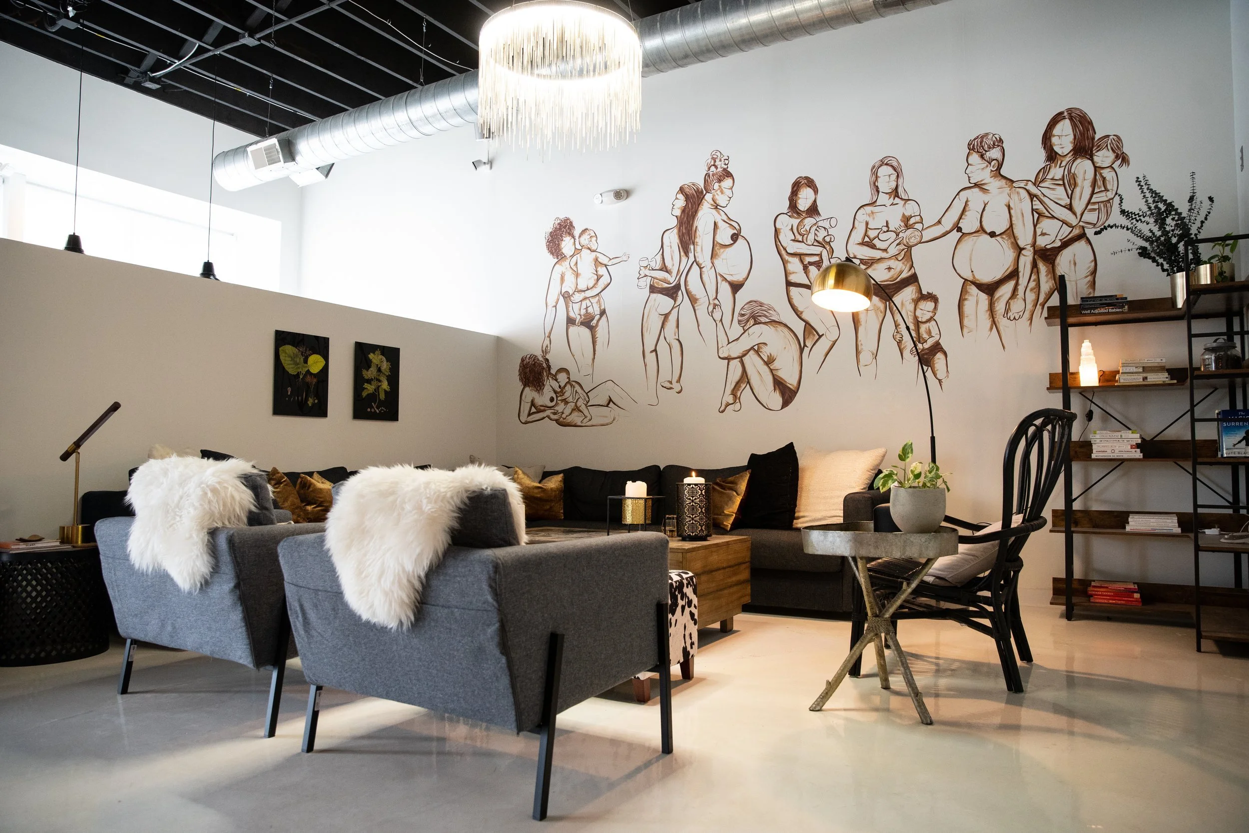 Modern living room with black sectional sofa decorated with white faux fur throws and pillows, a side table with a candle, a black metal bookshelf with books, potted plants, and decorative items, and a wall mural of line art illustrations of women an
