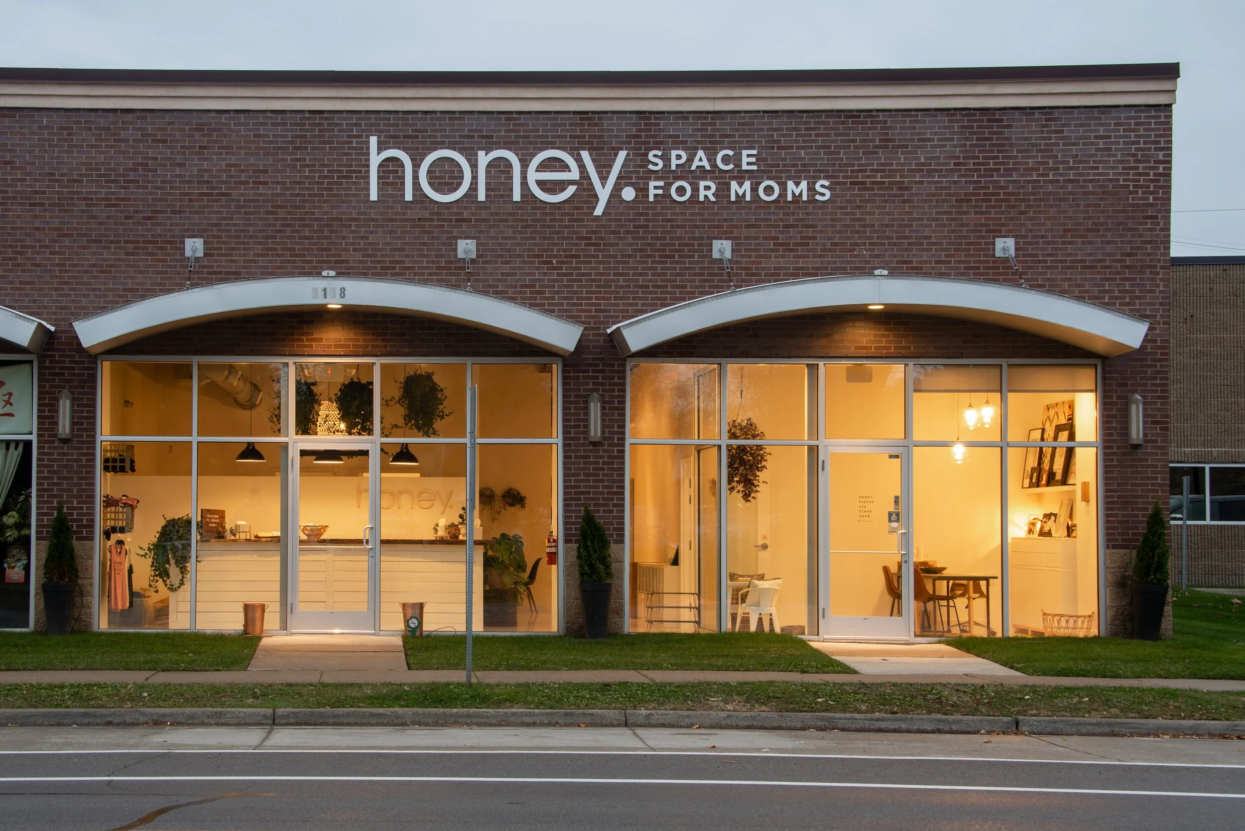  storefront with large glass windows and a sign on brick building reading "honey. SPACE FOR MOMS." The interior is warmly lit and decorated with plants and furniture.