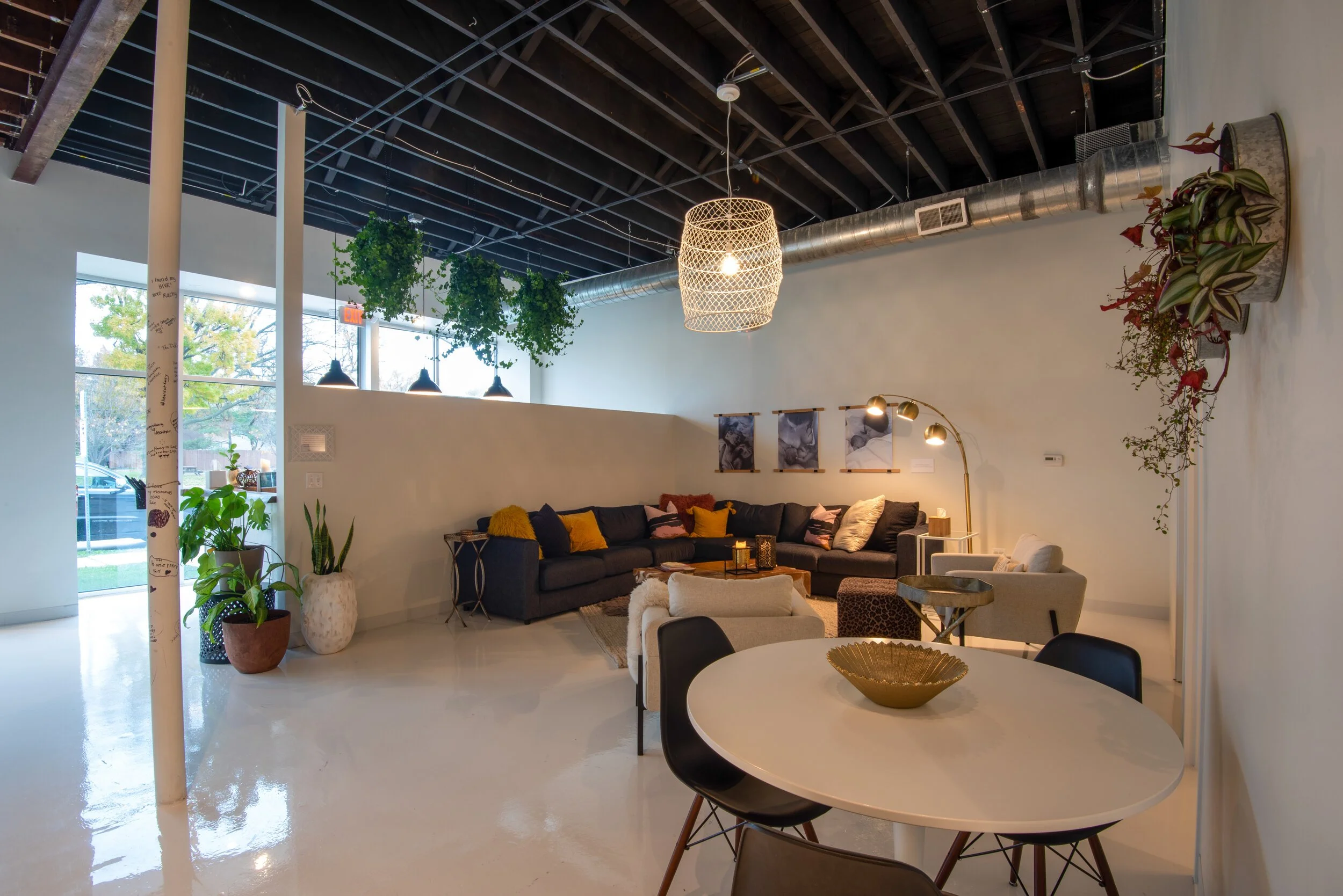 Modern lounge area with a black sectional sofa, white armchair, and round dining table. Decor includes hanging plants, wall art, and a woven pendant light, in an industrial-inspired space with exposed ductwork and a high ceiling.