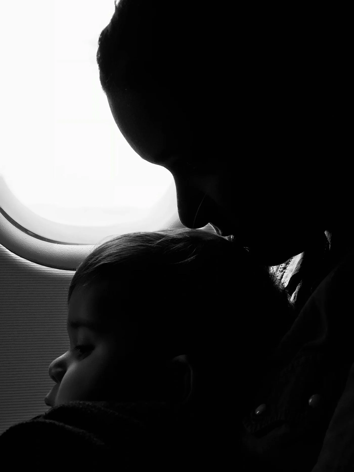 Silhouette of a man kissing a young girl on the forehead, both with closed eyes, near an airplane window.