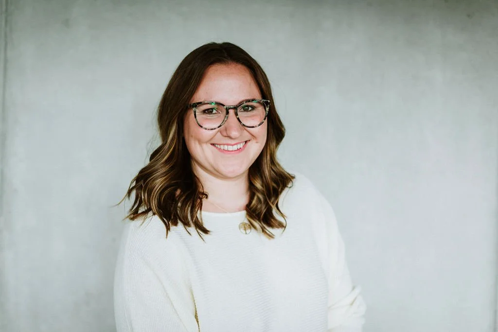 Eine lachende junge Frau mit braunen, gewellten Haaren und Brille trägt ein weißes Oberteil vor hellem, neutralem Hintergrund.