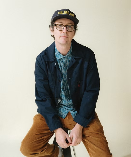 A man wearing glasses, a black cap with yellow text, a blue jacket, and brown pants sitting on a stool against a plain light-colored background.