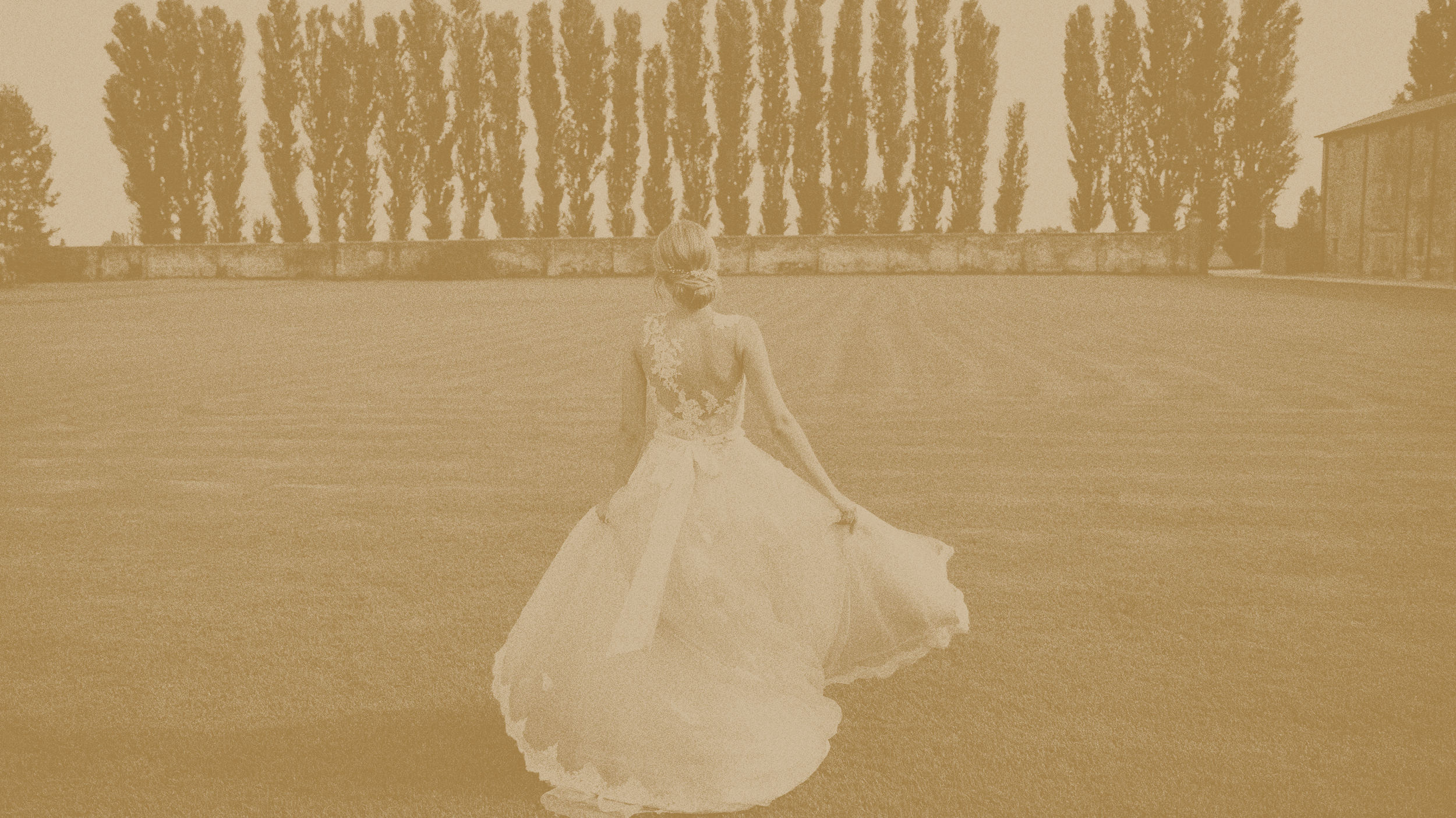 A woman in a wedding dress walking on a grassy field, with trees and a building in the background.