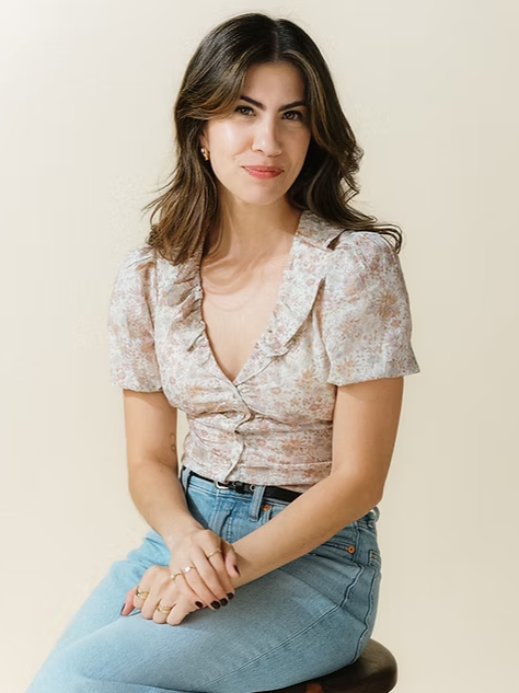 Woman with wavy brown hair wearing a floral blouse and blue jeans, sitting on a stool against a cream background.