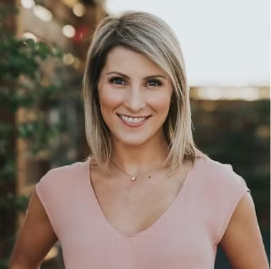 A smiling woman with shoulder-length blonde hair wearing a light pink top, standing outdoors near greenery.