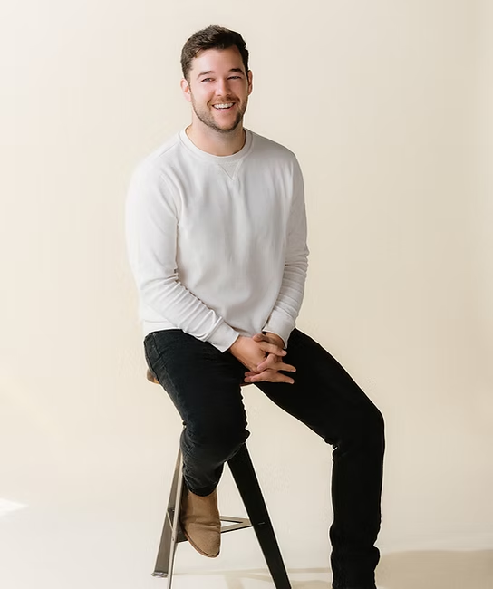 A man with short brown hair and a beard sitting on a stool, smiling, wearing a white long-sleeve shirt and black pants.