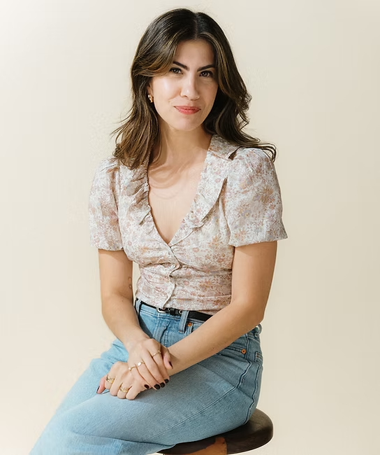 Woman with wavy brown hair wearing a floral blouse and light blue jeans, sitting on a stool against a plain background.