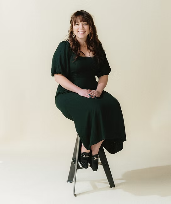 A woman with brown hair sitting on a black stool against a plain light beige background, smiling and wearing a dark green dress with black shoes.