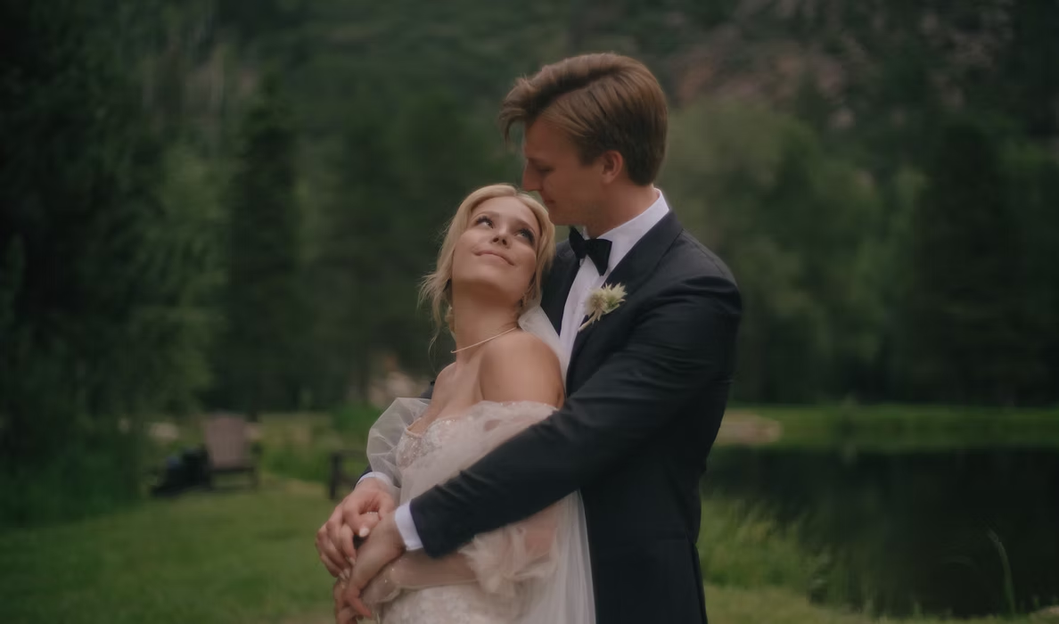 A bride and groom embrace outdoors by a pond, surrounded by greenery, during their wedding.