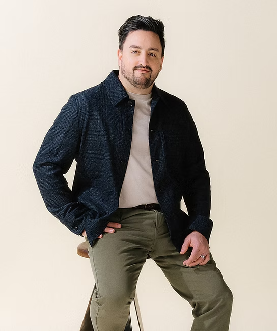 A man with dark hair and a beard, wearing a dark blue jacket, light gray t-shirt, and olive green pants, sitting on a wooden stool against a plain light background.