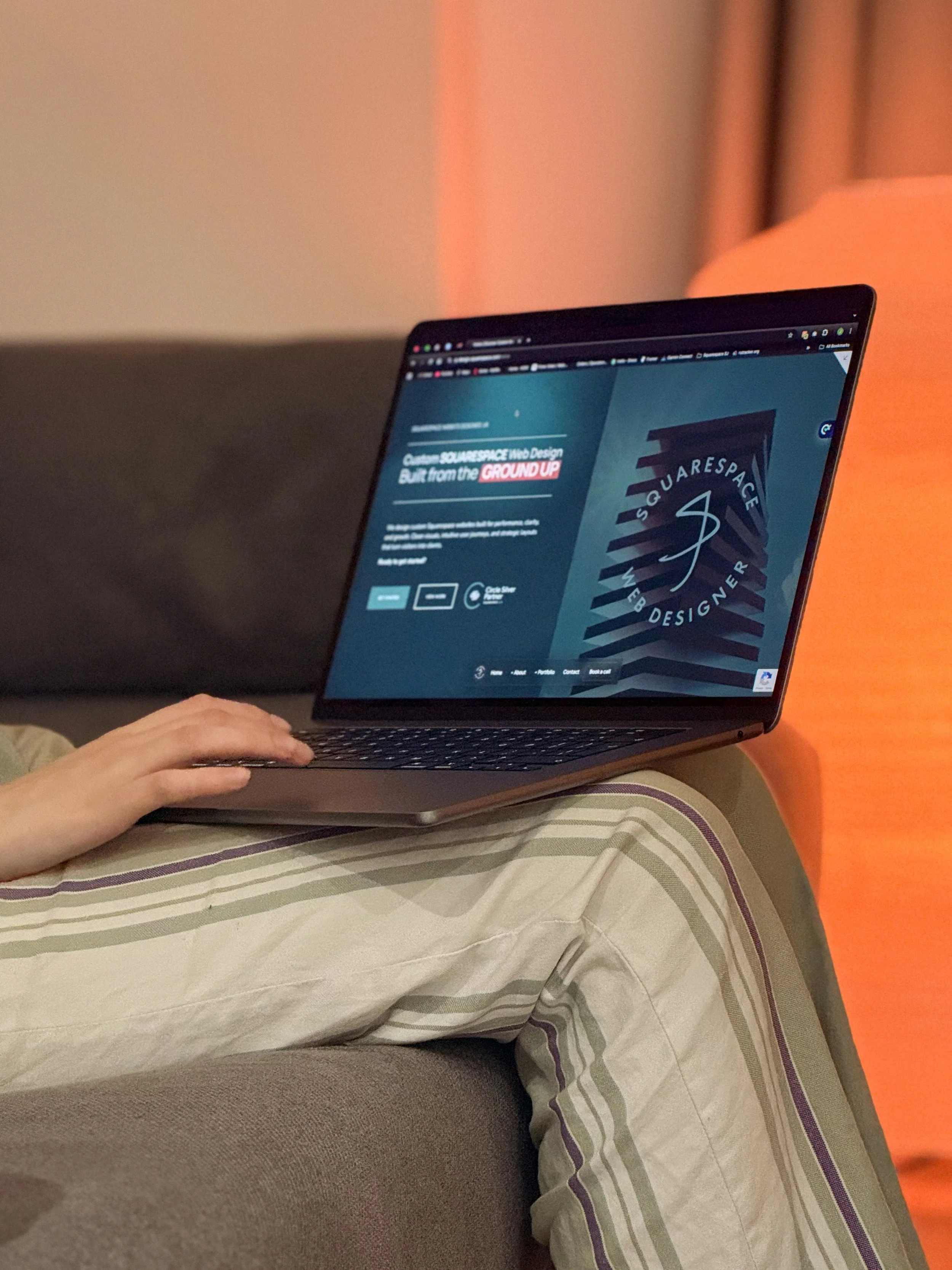 Person using a laptop on their lap, displaying a webpage about Squarespace web design, sitting on a couch with beige and striped pants, in a room with a dimly lit background and orange furniture.