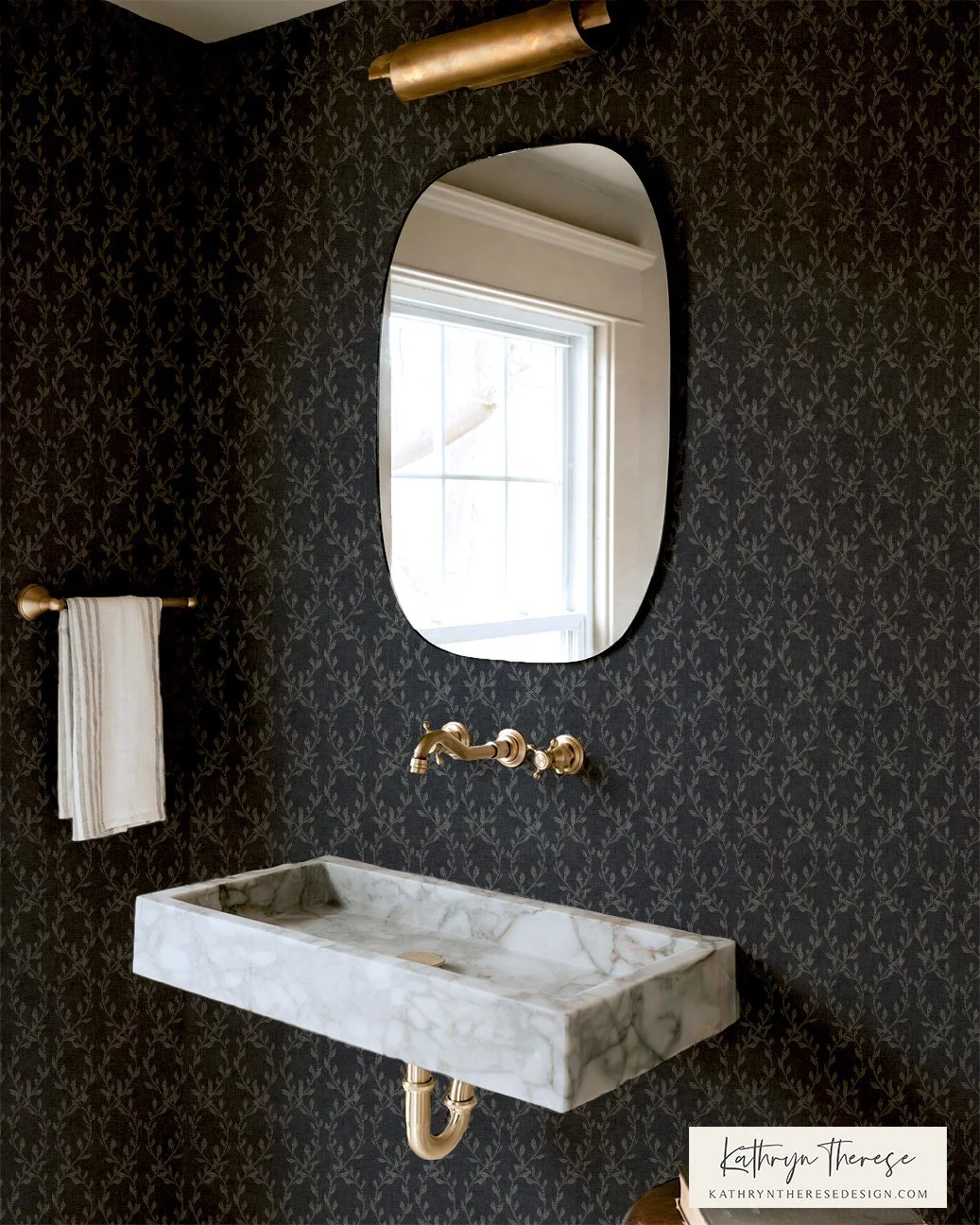 Bathroom with black patterned wallpaper, oval mirror, marble wall-mounted sink, gold fixtures, and a window reflected in the mirror.
