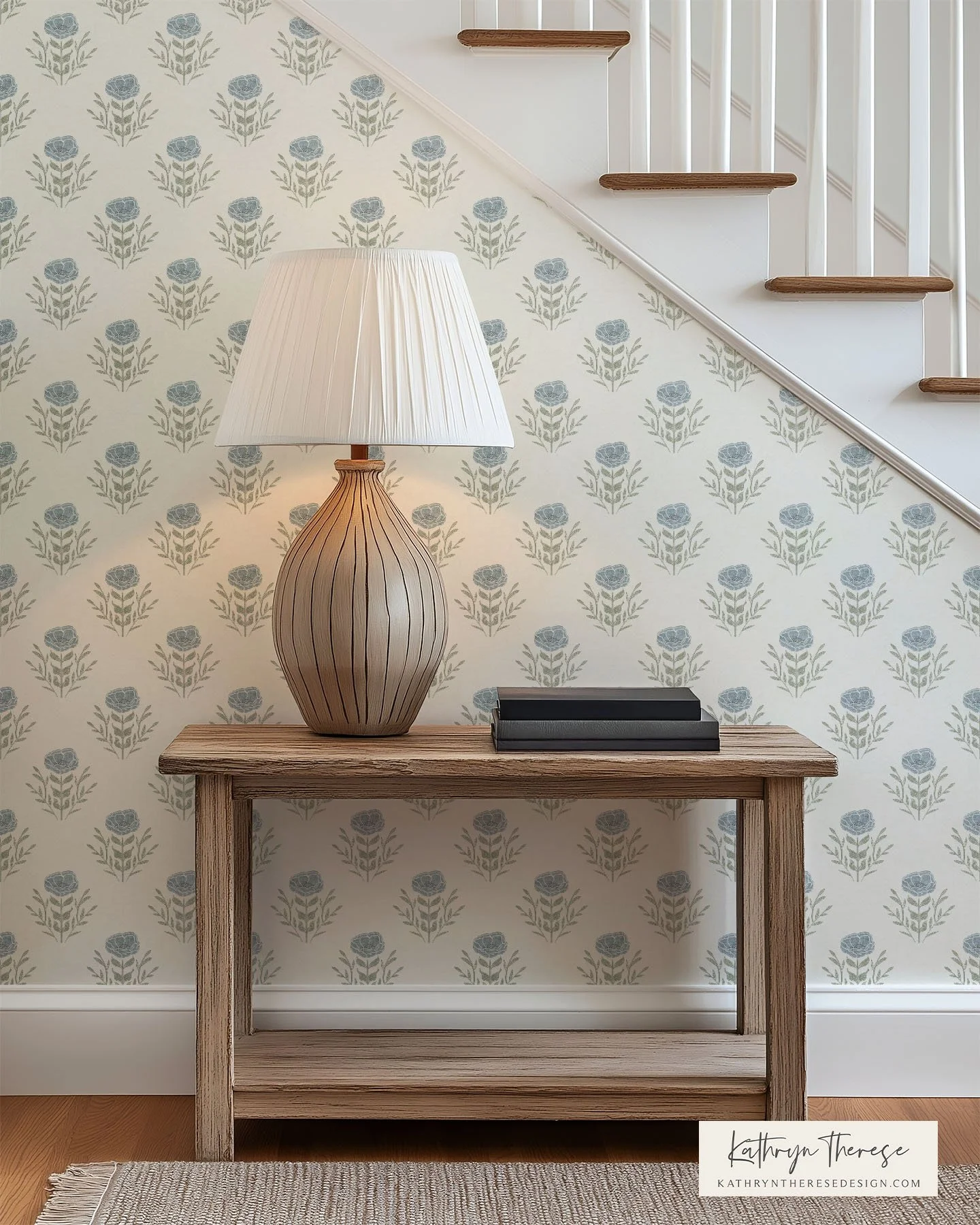 A wooden console table with a large beige striped ceramic lamp and three black books on top, against a wallpapered wall with a floral pattern, next to a staircase with white railing and wooden stair treads.