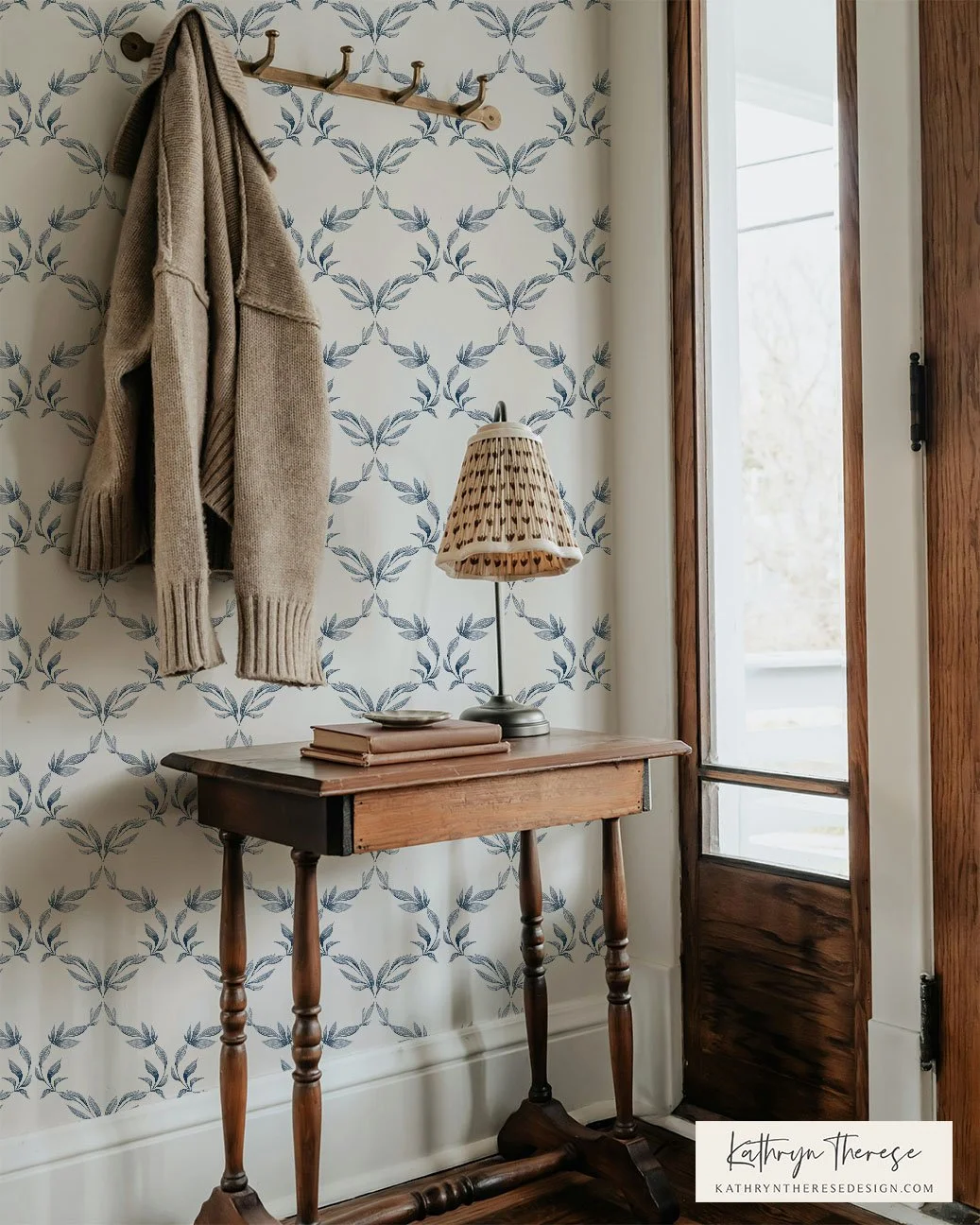 A cozy entryway corner with a wallpapered wall featuring a blue leafy pattern, a wooden coat rack with hooks holding a beige sweater, a small wooden side table with turned legs, a beige and brown lamp with a woven shade, and a window with a wooden fr