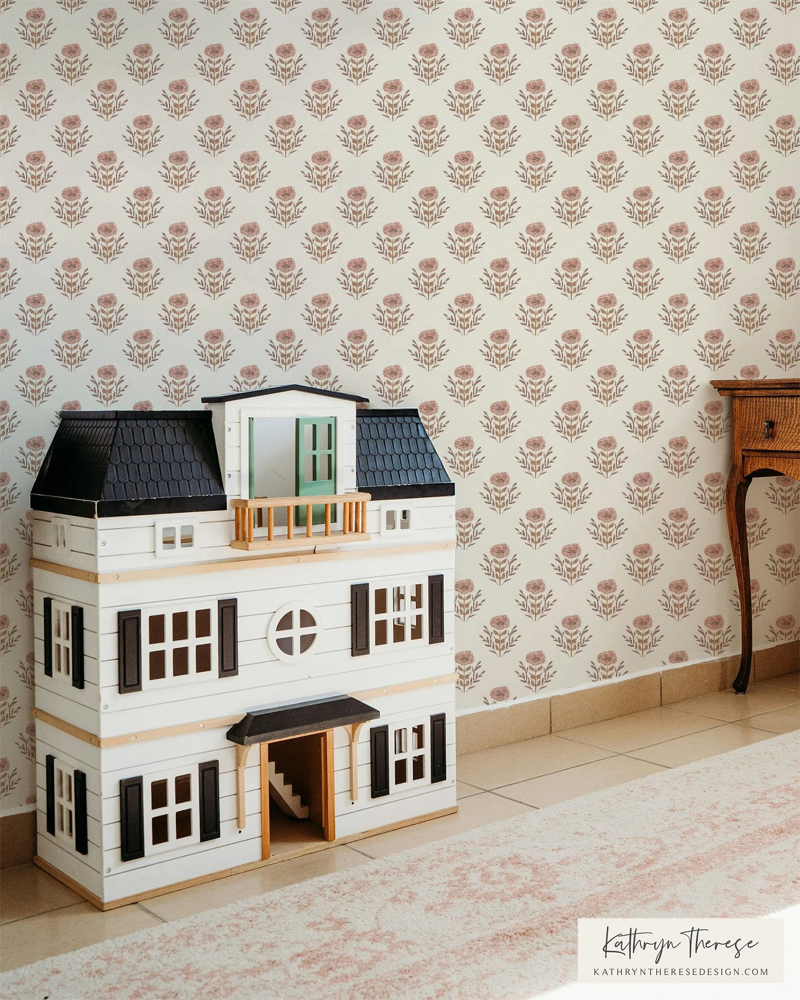 A dollhouse with multiple stories and black window shutters, placed against a wallpapered wall with a floral pattern, next to a small wooden side table.