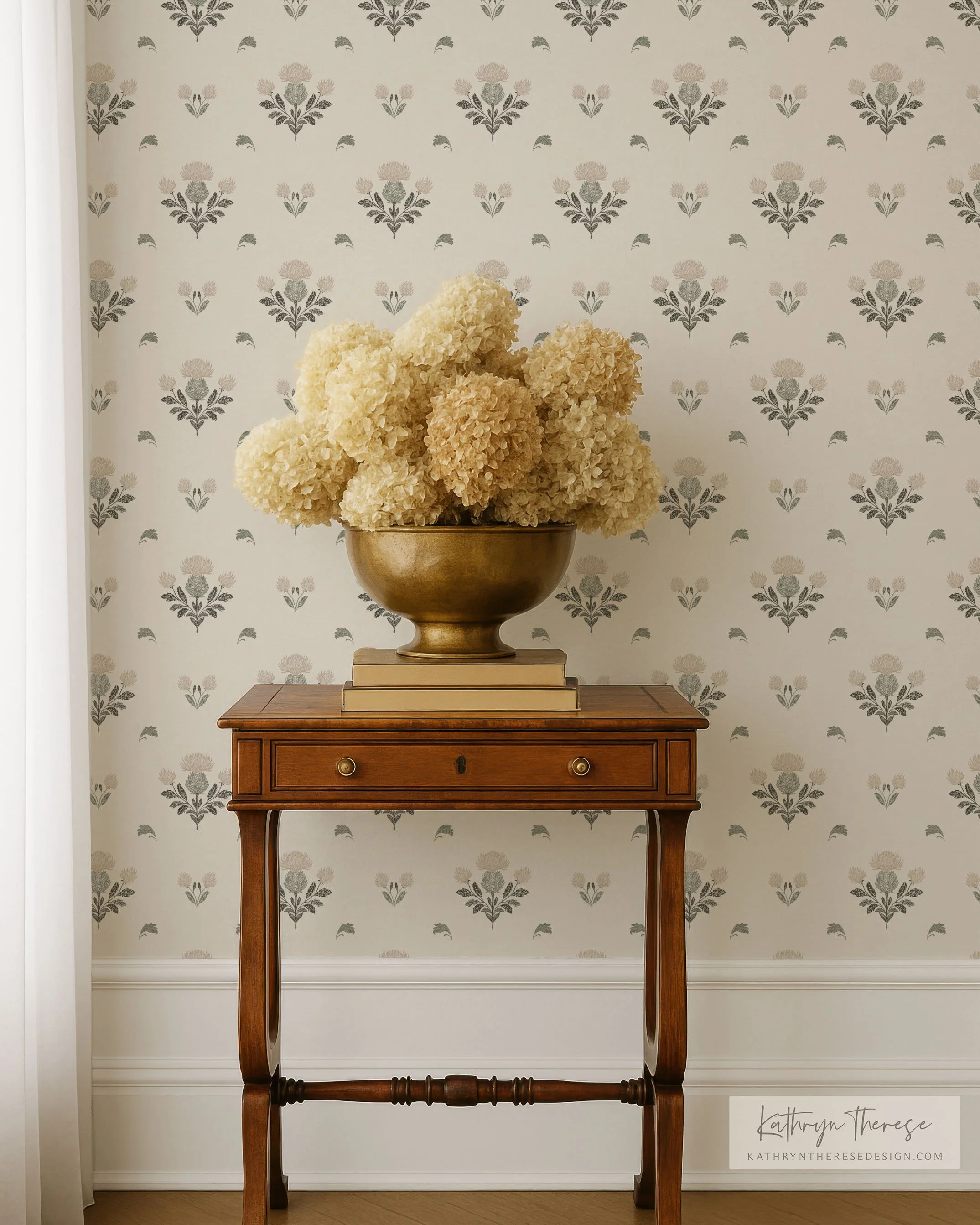 A wooden table with a drawer, holding a metallic bowl filled with cream-colored hydrangea flowers, against a floral wallpaper background.