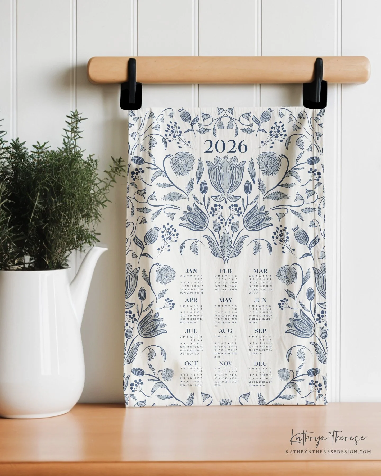 2026 wall calendar with blue floral design hanging on a wooden rod, placed on a table next to a white watering can with green plants inside, and a white paneled wall background.