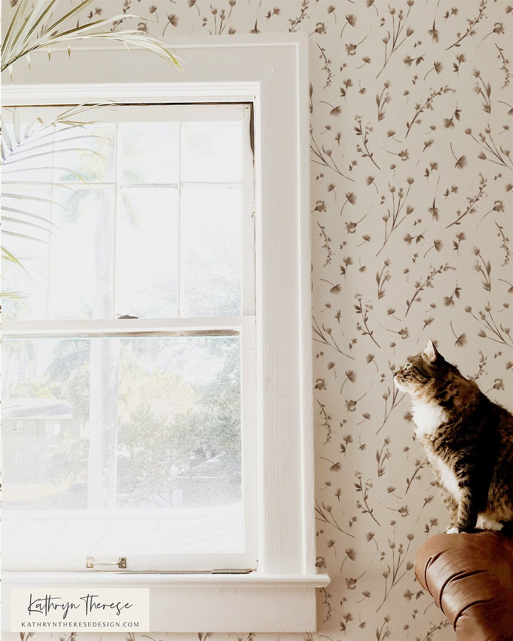 A cat sitting on a brown armrest looking out of a white-framed window with sheer curtains, against a wall with beige floral wallpaper, and a palm leaf partially visible in the top left corner.