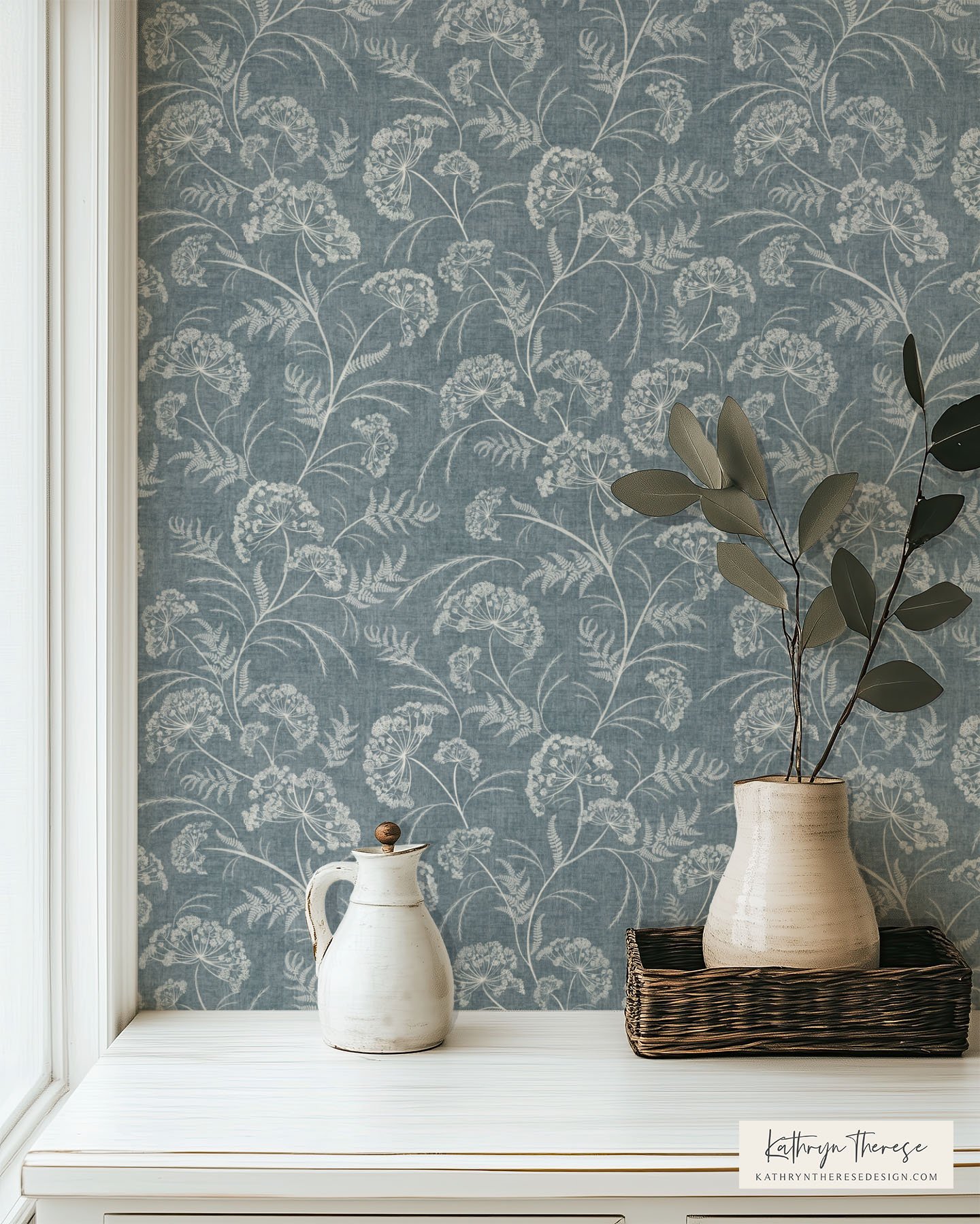 A white pitcher with a knob on top, a white ceramic vase with large dark green leaves, on a white surface near a window, with light blue wallpaper featuring a white floral and fern pattern.