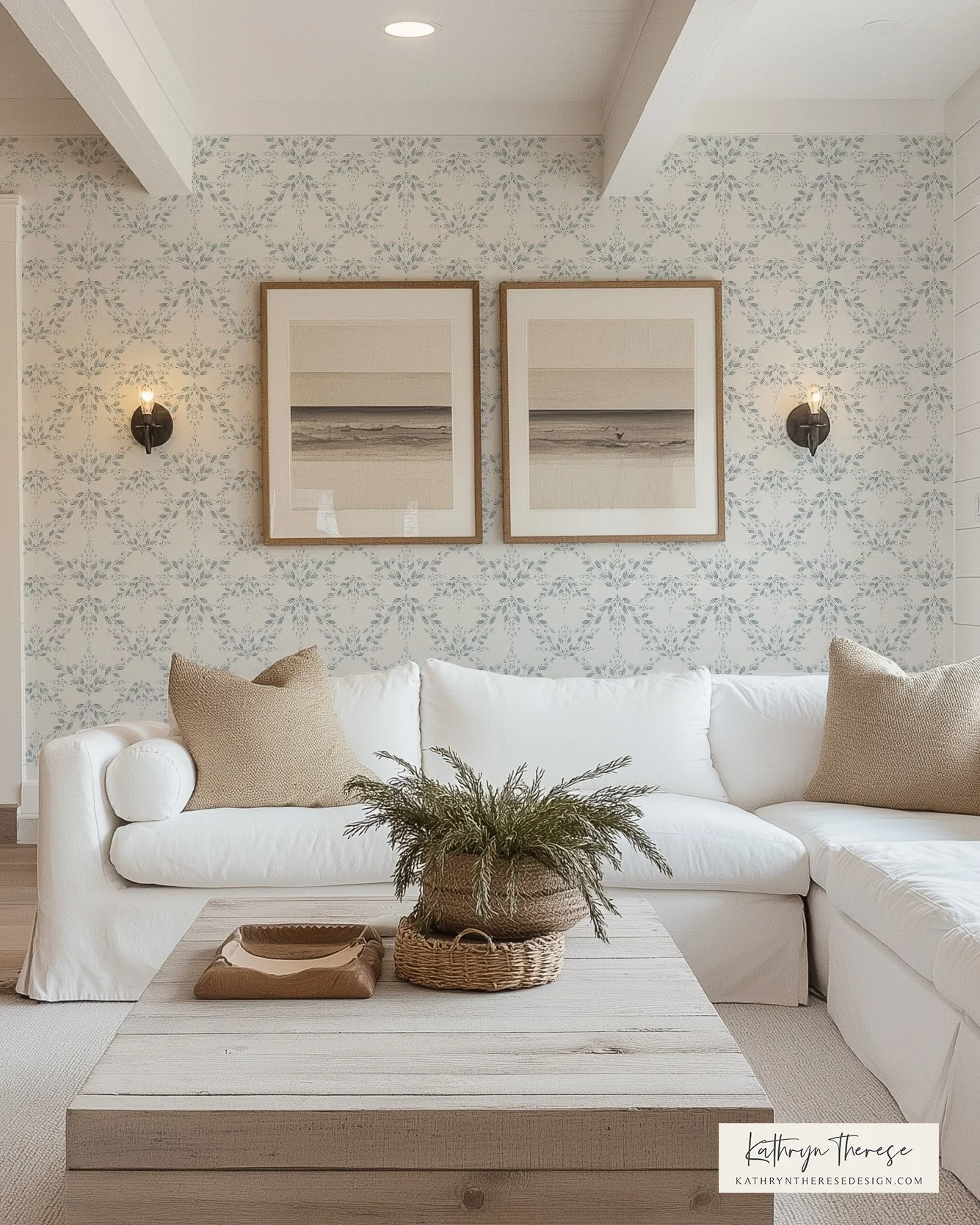 Living room with white sectional sofa, beige pillows, wooden coffee table with green plant in a wicker basket, patterned wallpaper, two framed beach photographs, and wall sconces.