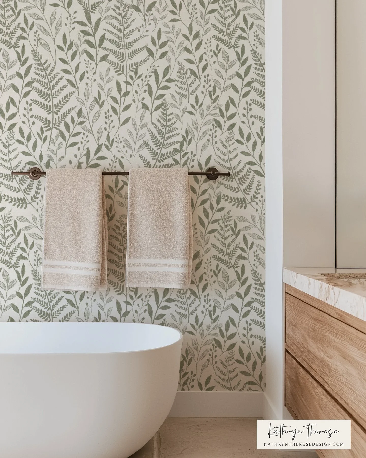 Modern bathroom with a white freestanding bathtub, beige towels hanging on a rod, light wood vanity, marble countertop, and botanical leaf-patterned wallpaper.