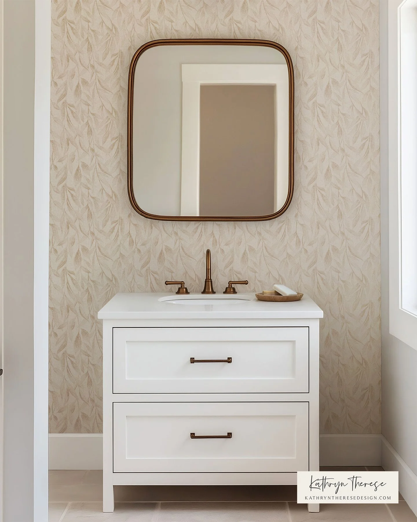 White bathroom vanity with two drawers, bronze handles, and a bronze faucet, above beige patterned wallpaper, with a square bronze-trimmed mirror, and a soap dish on the right side of the sink.