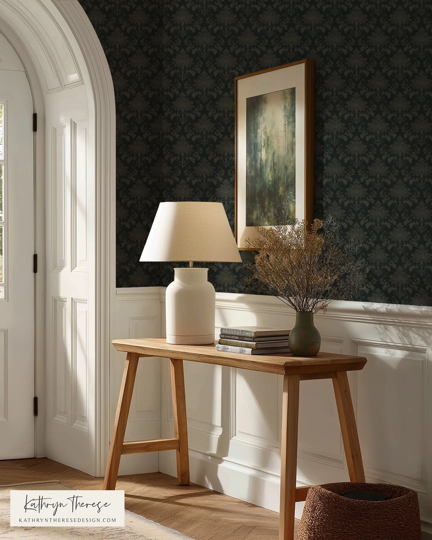 A hallway corner with a wooden console table, white table lamp, a stack of books, and a vase with dried branches. Art hanging on a dark patterned wall and a door with decorative molding are visible.
