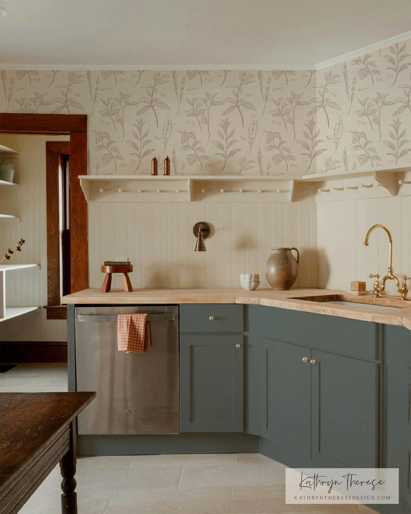 A cozy kitchen corner with a beige patterned wallpaper, a white wooden backsplash, and light blue cabinets. The countertop has decorative items including a large jar, a small bowl, and a stand. There is a brass faucet and a wooden window frame.