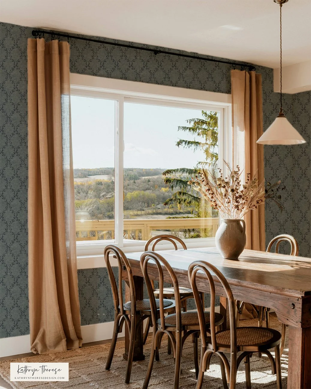 A dining room with a large window revealing a scenic landscape with trees and a cloudy sky. The room features a wooden dining table, six wooden chairs, a white vase with dried flowers, patterned wallpaper, beige curtains, and a hanging light fixture.