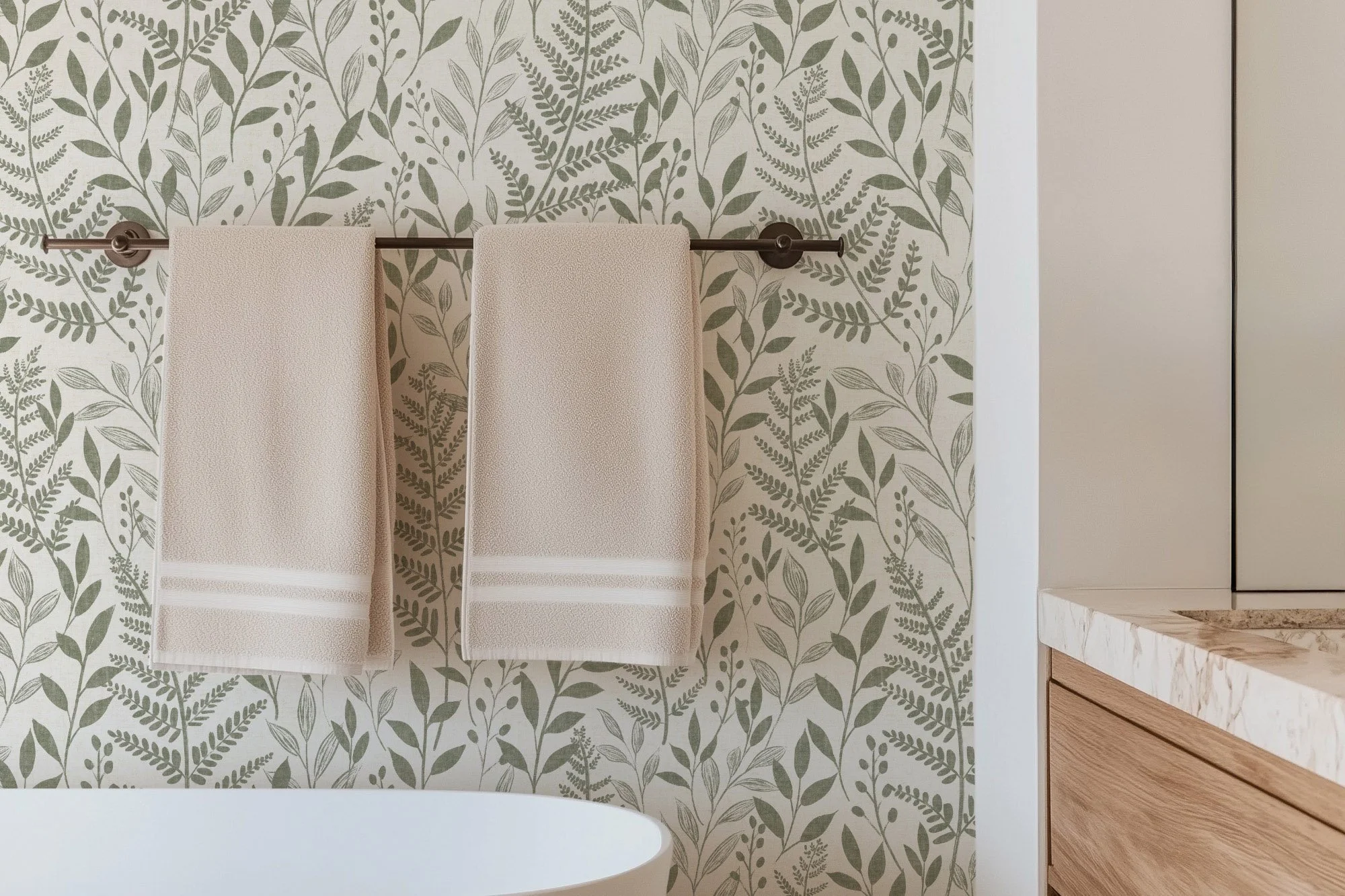 Two beige towels hanging on a black towel rod against a wall with leafy botanical wallpaper in shades of green and beige, next to a cream-colored cabinet with a marble countertop.