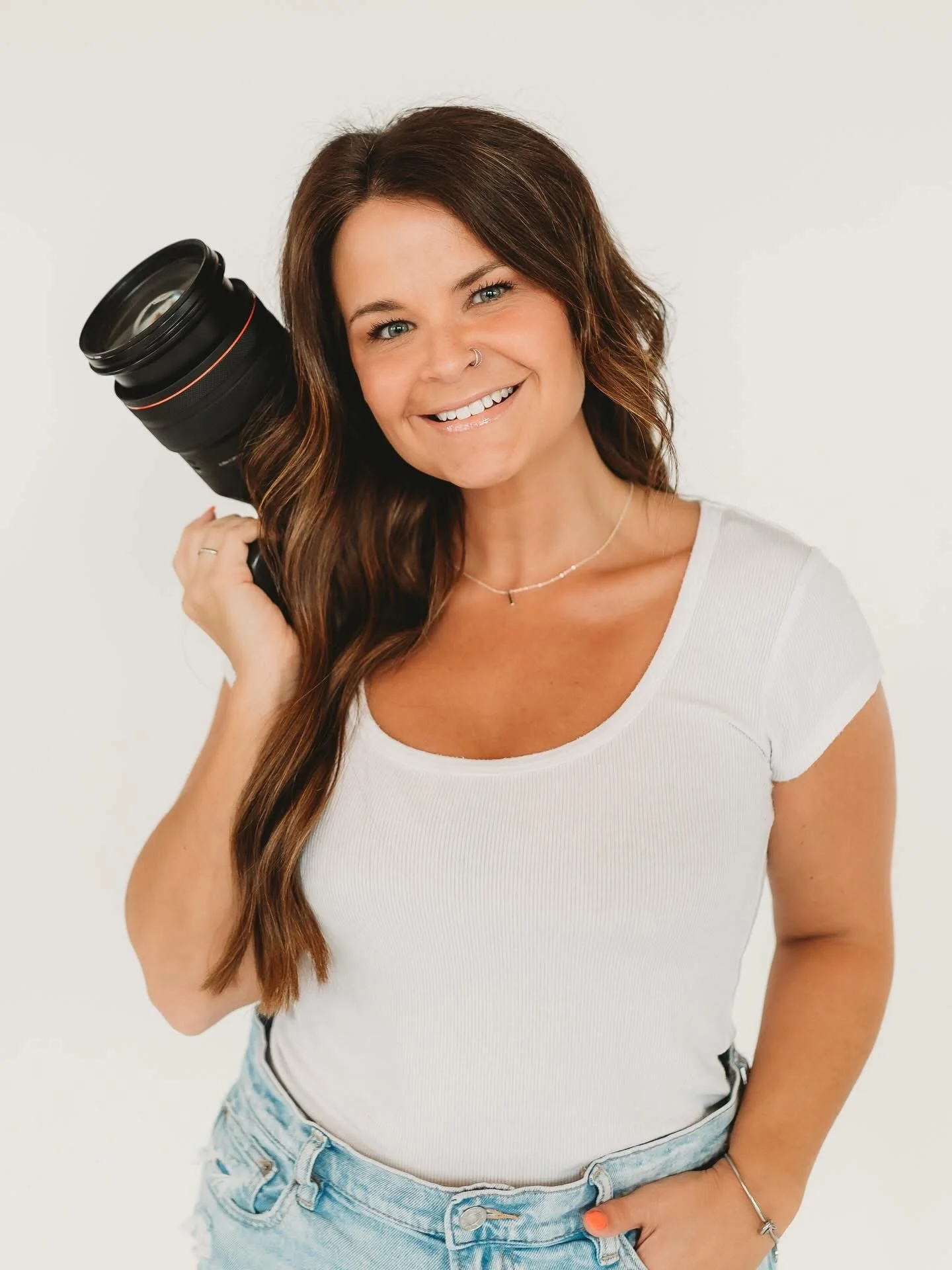 Smiling woman with long brown hair holding a camera lens over her shoulder, wearing a white t-shirt and light blue jeans.