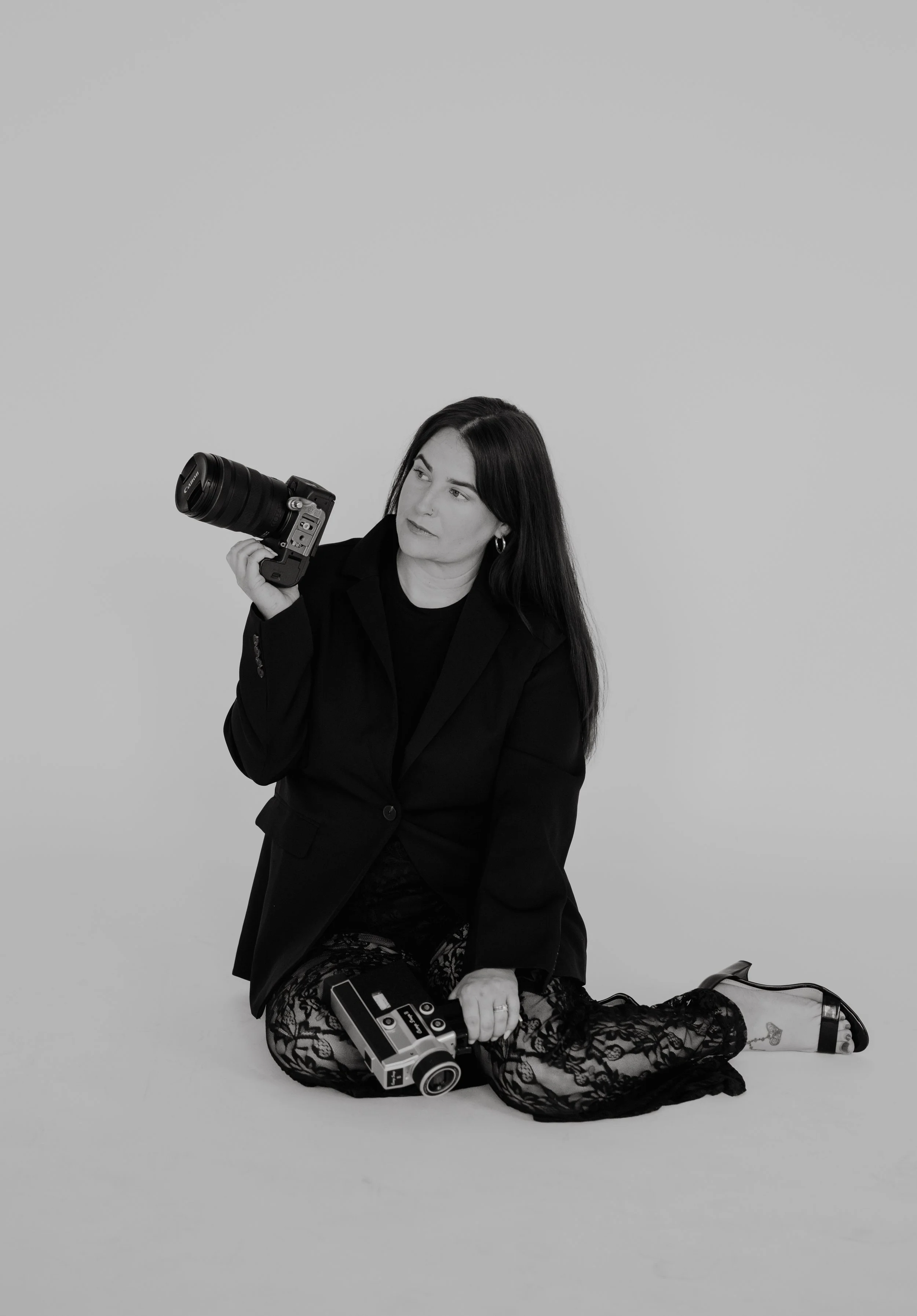 Black and white photo of a woman sitting on the floor, holding a camera and a camera case, wearing a blazer and lace pants, with a plain background.