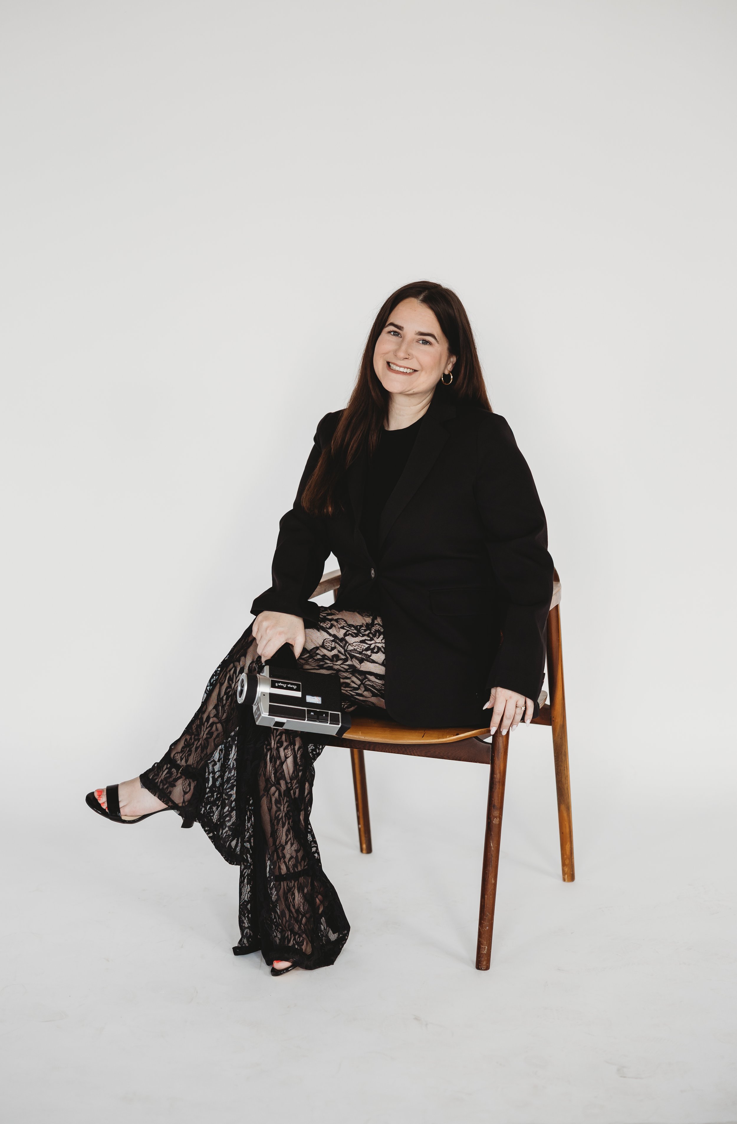 Woman with long brunette hair, wearing a black blazer and lace pants, sitting on a wooden chair, smiling, holding a vintage camera, against a plain white background.