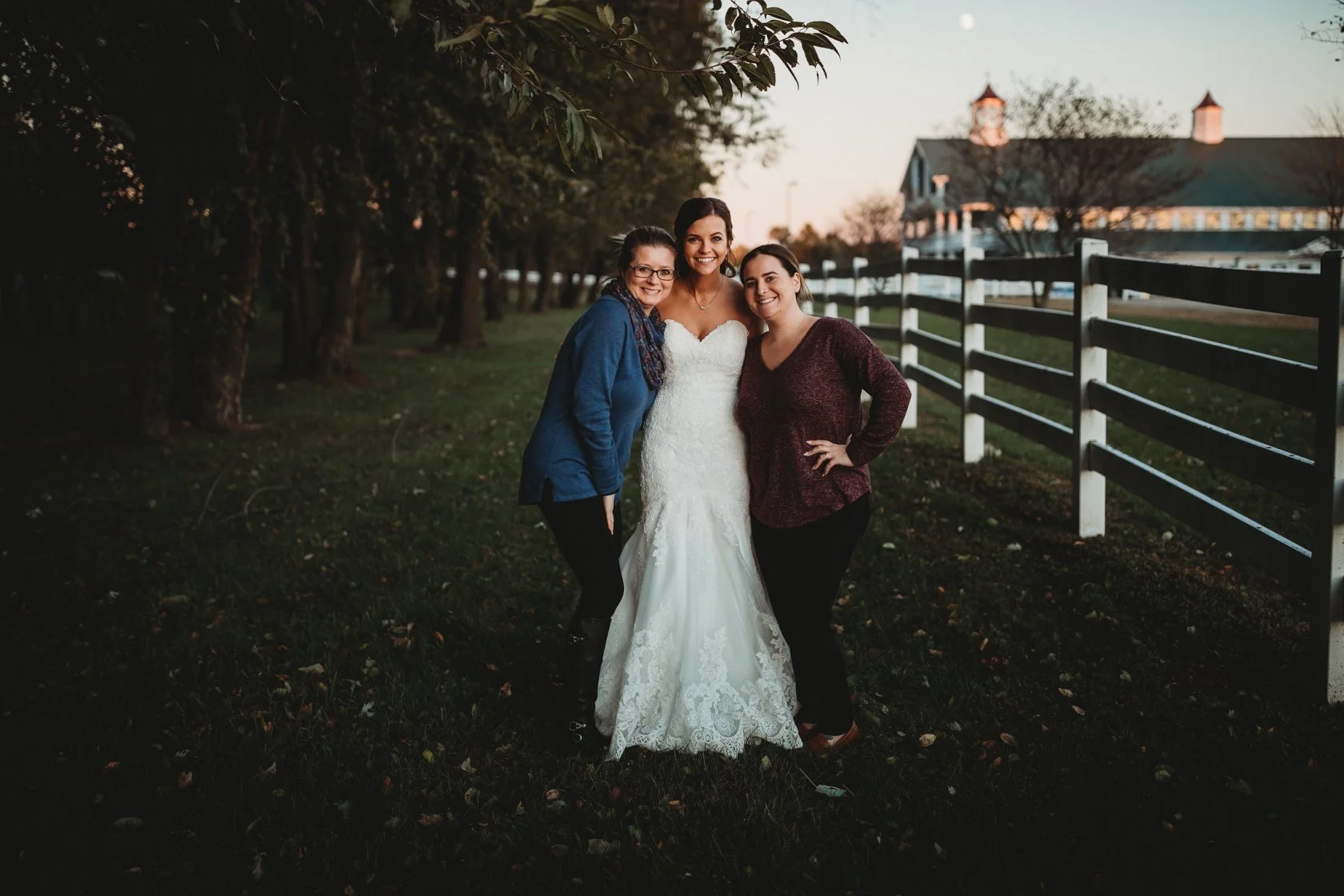 Three women, including a bride in a white wedding gown, standing outdoors on grass at sunset, smiling and posing for a photo.